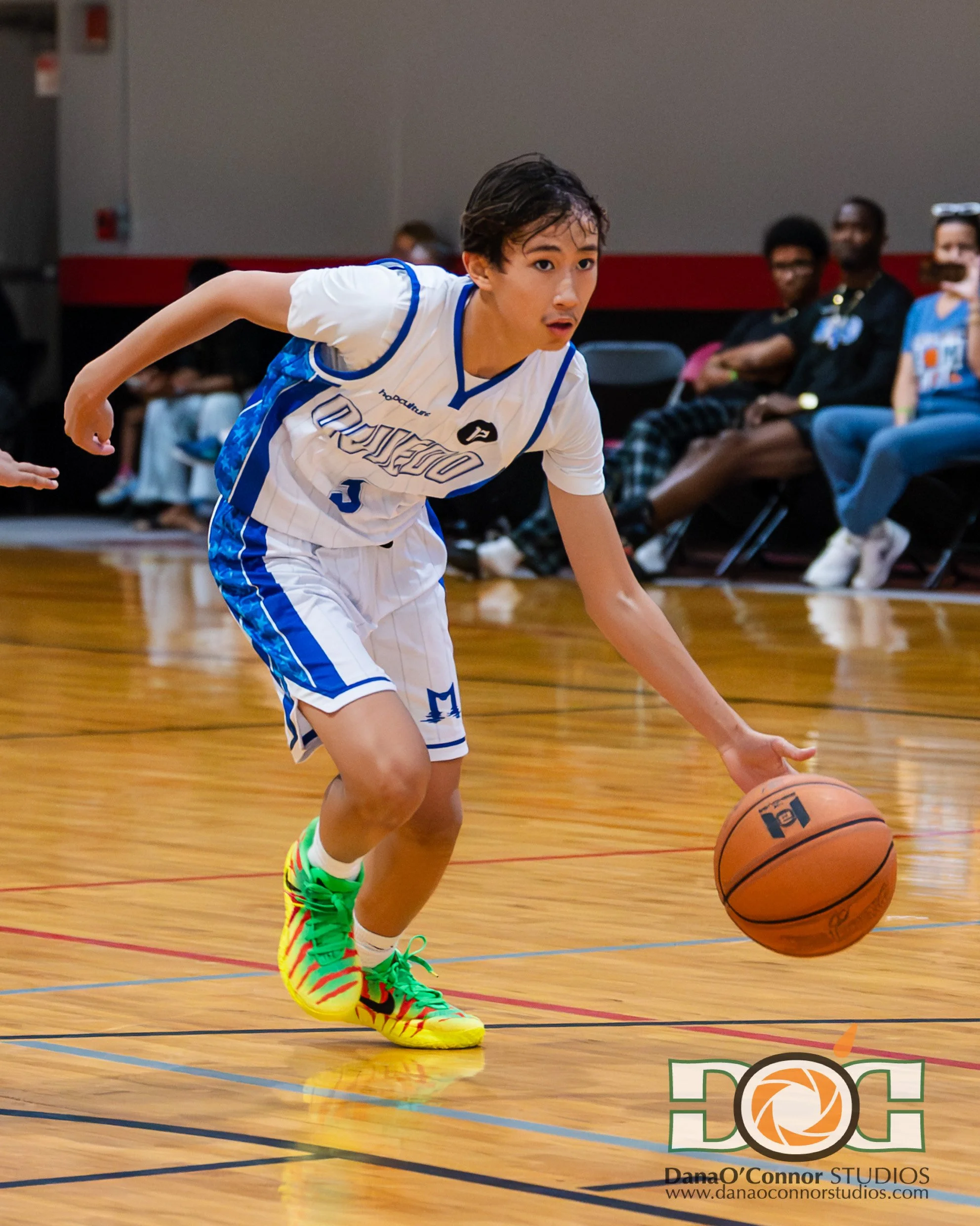 A young male basketball player in a white and blue uniform dribbling a basketball on an indoor court, with spectators seated in the background.