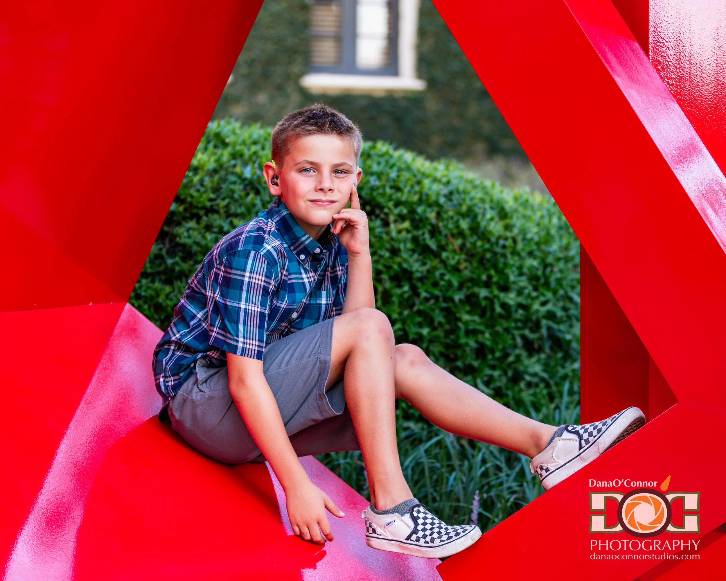 A young boy with short hair, wearing a blue plaid shirt and gray shorts, sitting on a pink slide with a red structure, outdoors with green bushes and trees in the background.
