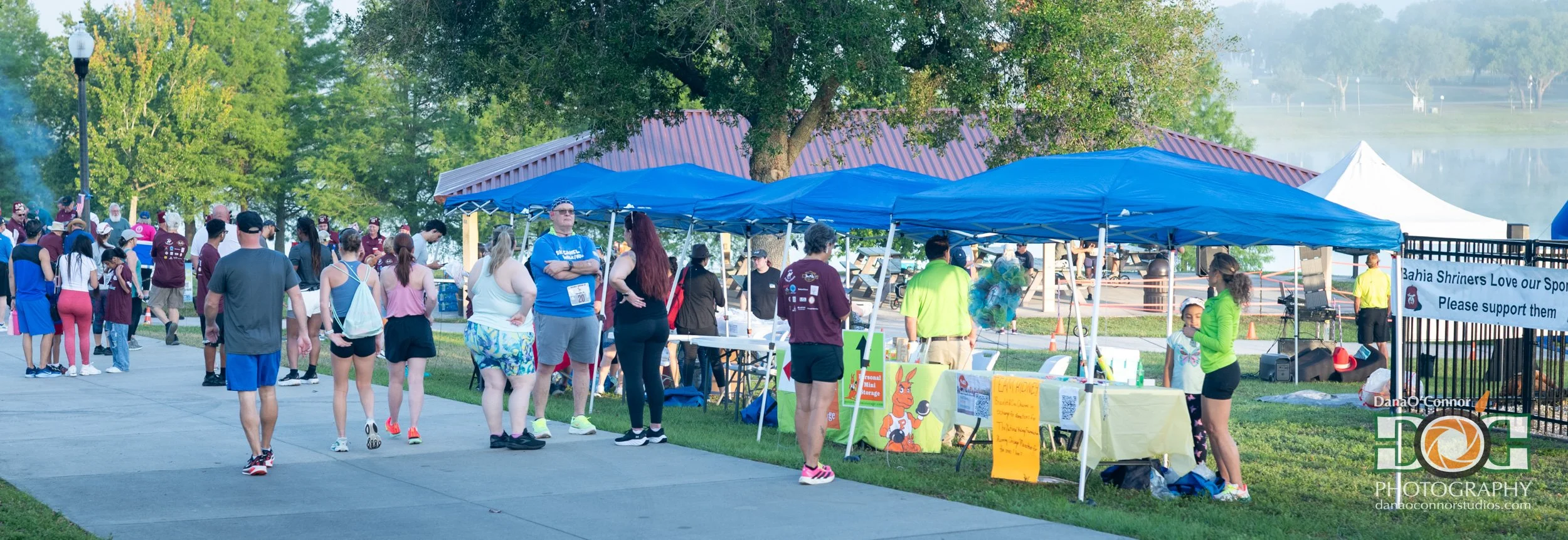 People gathered at an outdoor event with tents, tables, and a lake in the background. Participants are walking or standing, some wearing athletic clothing. There are signs and banners, including one that reads 'Bahia Shriners Love our Sport. Please s