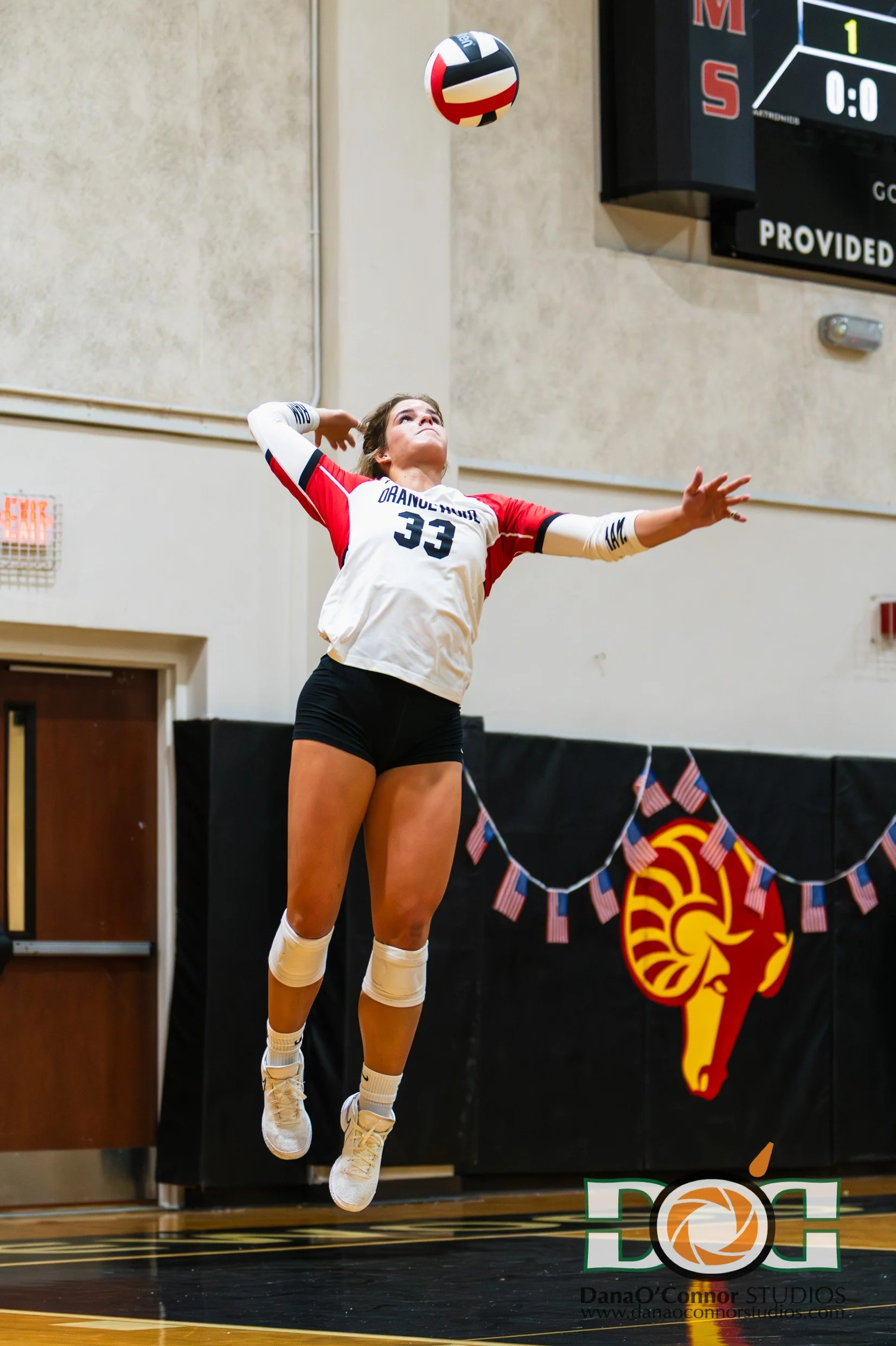 A female volleyball player wearing a white jersey with the number 33, with red and black accents, is jumping in mid-air to hit a volleyball during a game. She is wearing black shorts, white knee pads, and white athletic shoes. The background shows a 