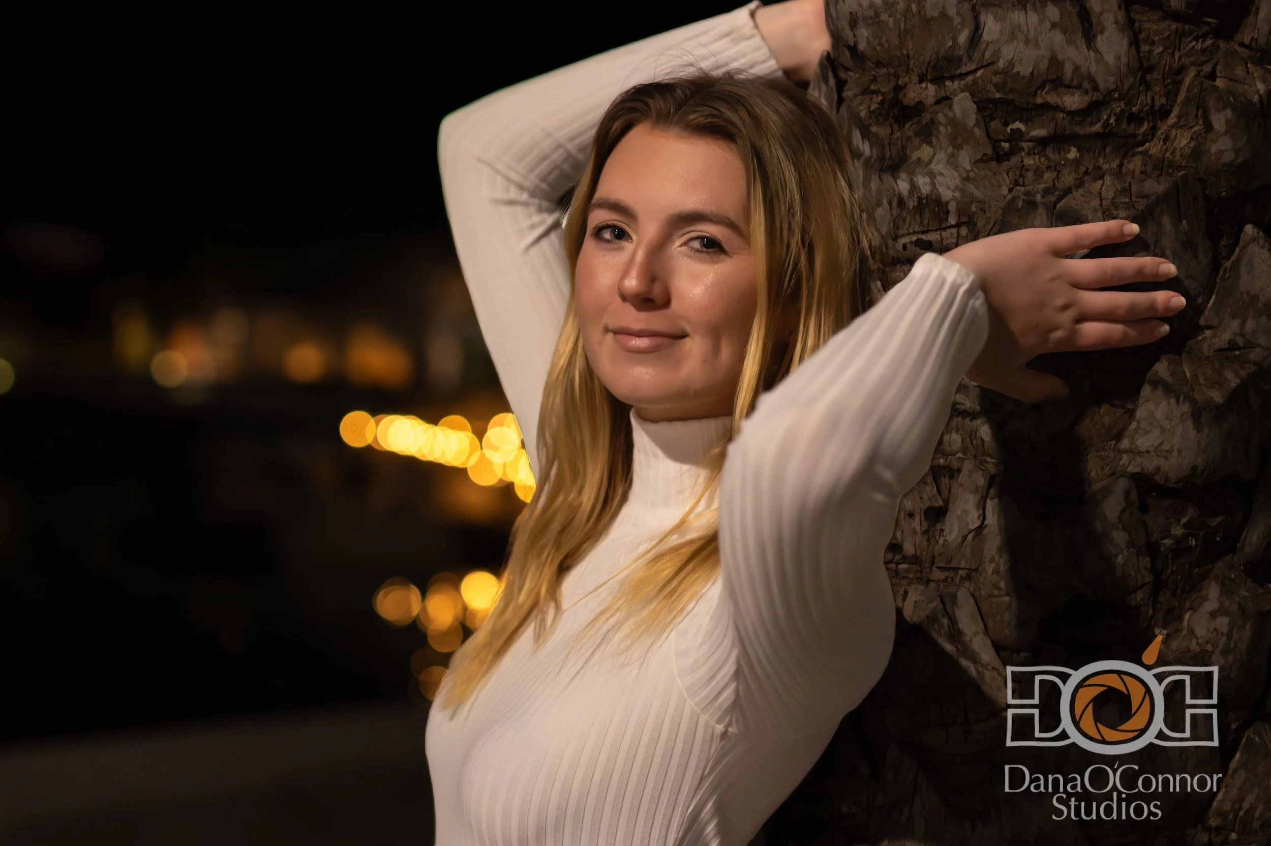 A young woman with long blonde hair leaning against a tree at night, smiling softly. She is wearing a white long-sleeve shirt and is illuminated by warm ambient lighting.