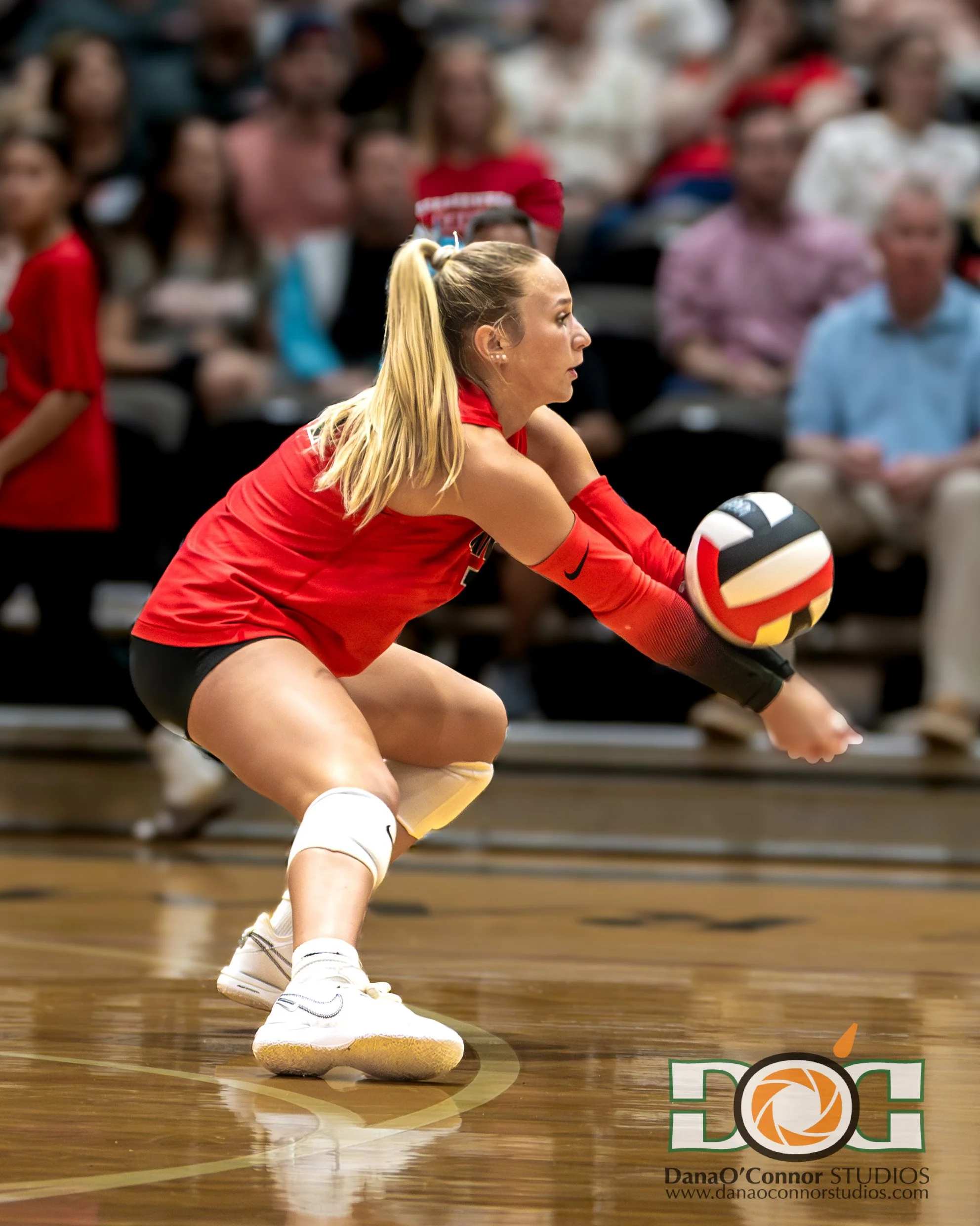 A female volleyball player in red uniform and white shoes crouches with her arms extended, preparing to hit the ball during a game on a wooden court with spectators in the background.