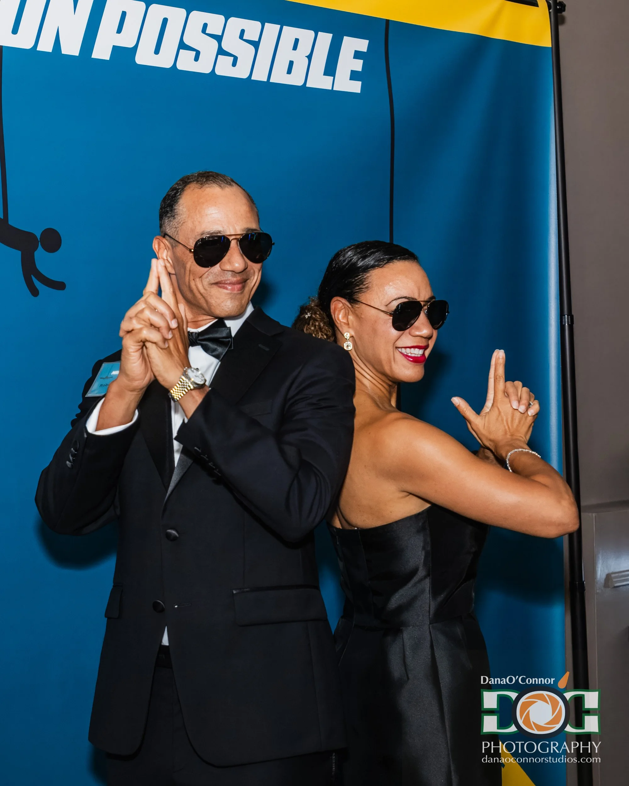 Two people in formal attire posing with sunglasses in front of a blue and yellow backdrop with the word 'IMPOSSIBLE' at the top and a logo at the bottom right corner.