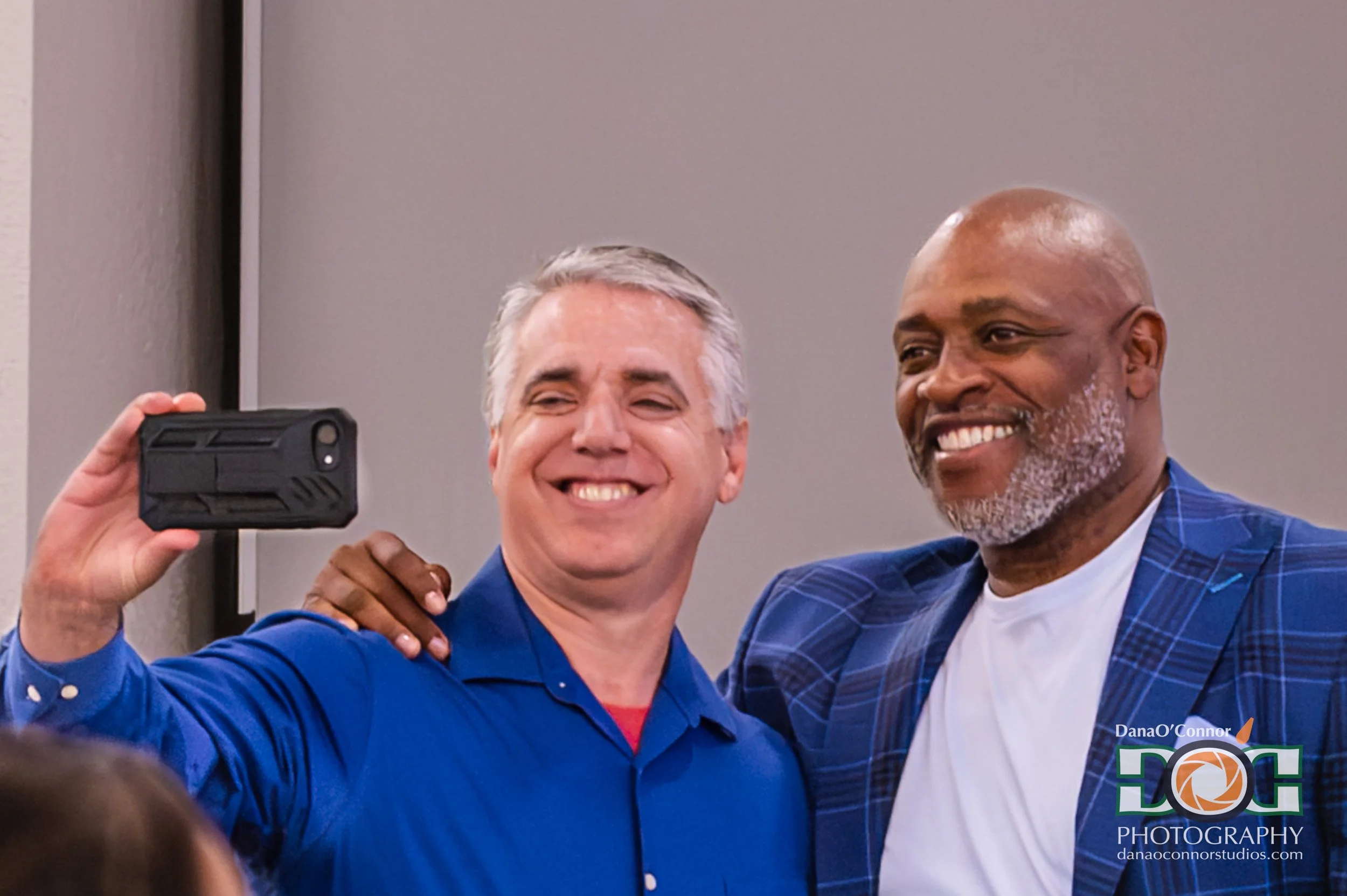 Two men taking a selfie, one with gray hair and the other with a bald head and facial hair, smiling and wearing blue suits, in a professional setting.