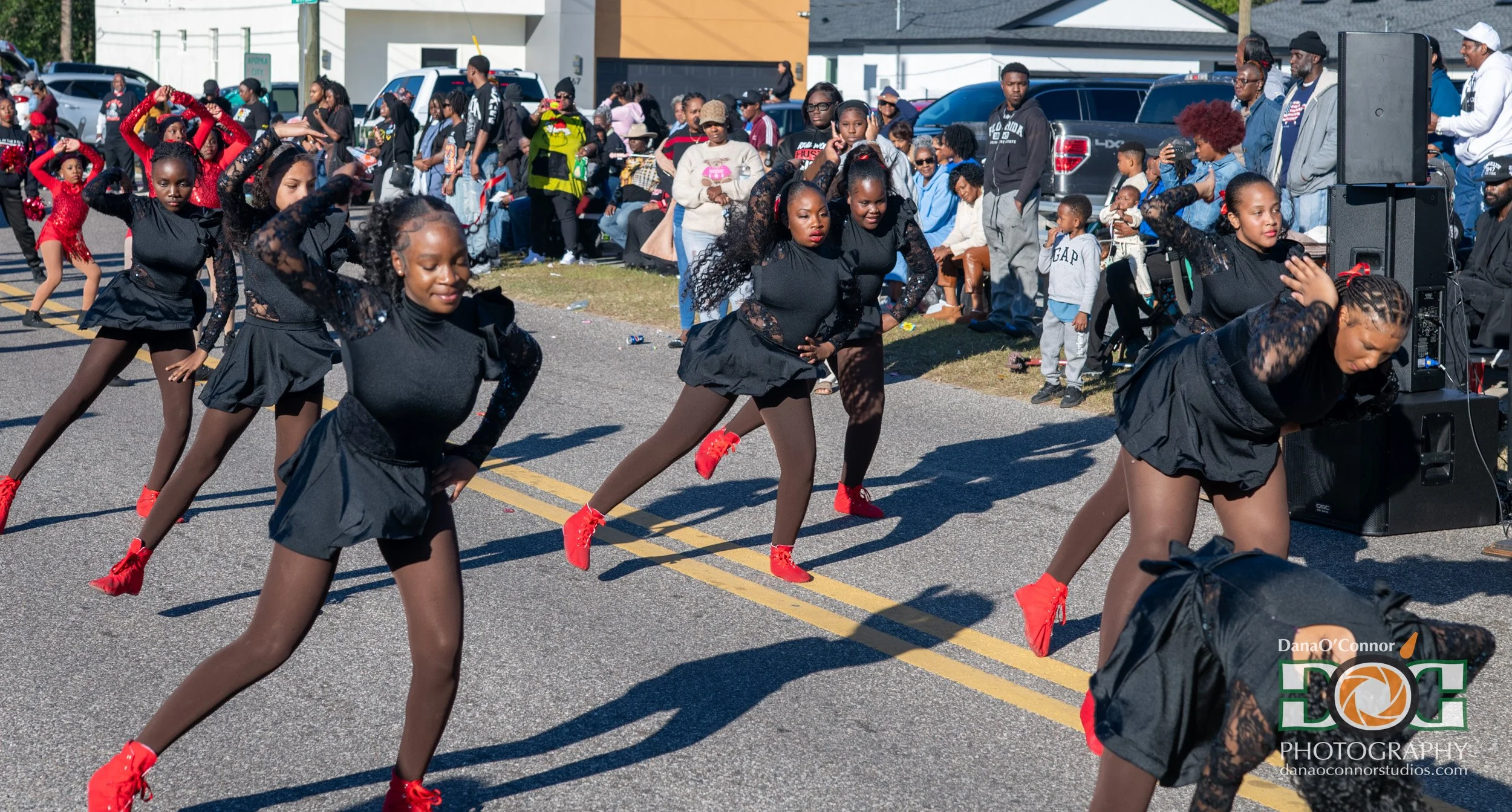 A group of young girls performing a dance routine in matching black outfits and red shoes on a city street during an outdoor event, with an audience of spectators sitting and standing behind barricades and parked cars.