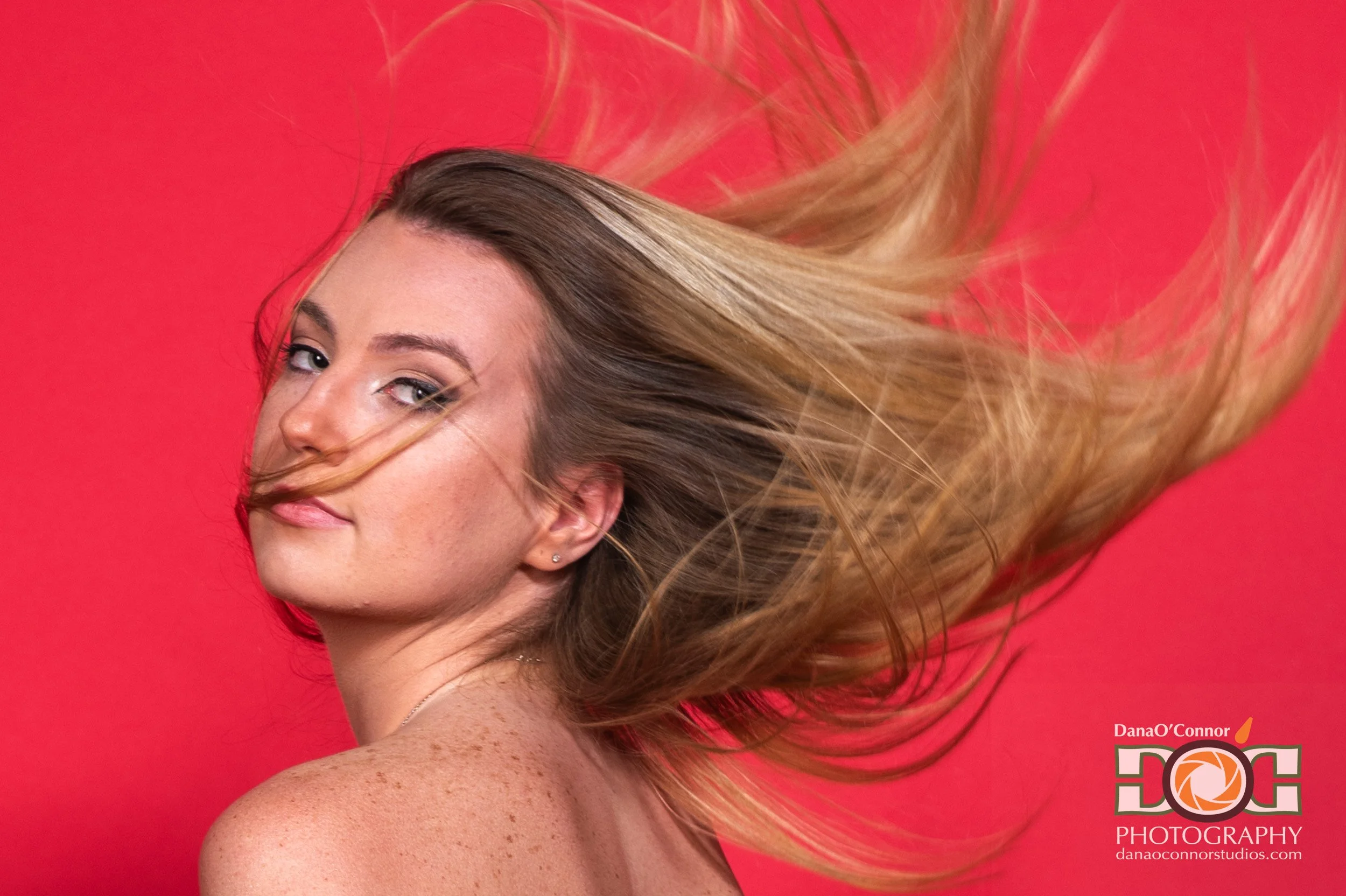 A woman with long, flowing blonde hair, wearing makeup, looking over her shoulder, against a red background. She has a small earring and is slightly smiling.