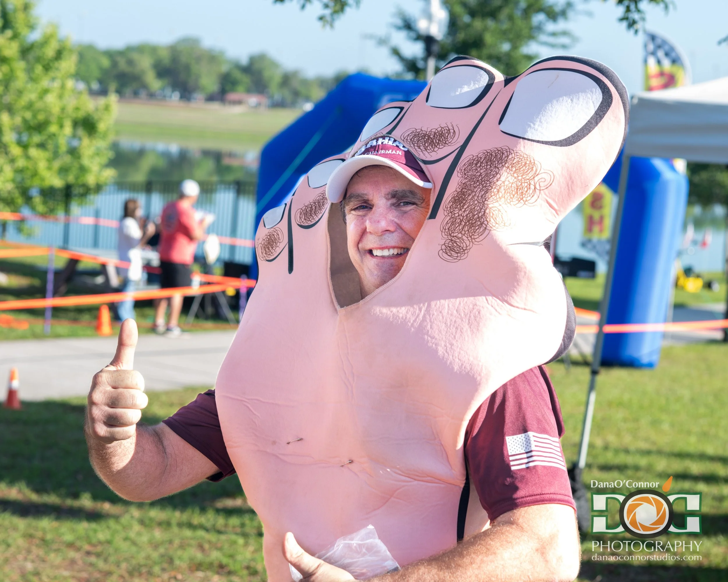 A man smiling and giving a thumbs-up wearing a costume that resembles a cartoon pig, outside at a park or event with trees and tents in the background.