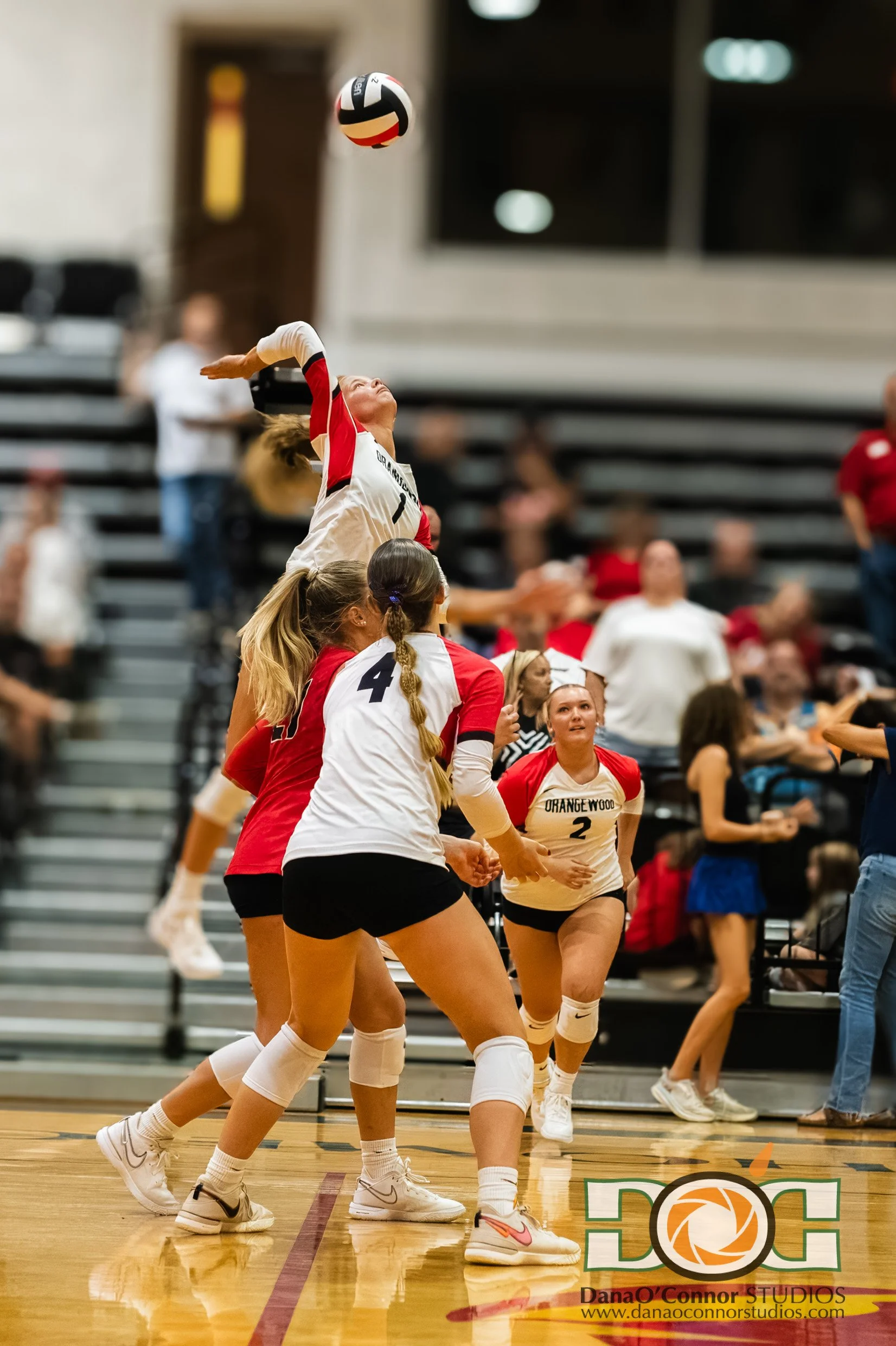 A group of young women playing volleyball indoors, jumping and reaching for the ball. They are wearing uniforms with numbers, and some are wearing knee pads and white sneakers. Spectators are seen sitting in the background.