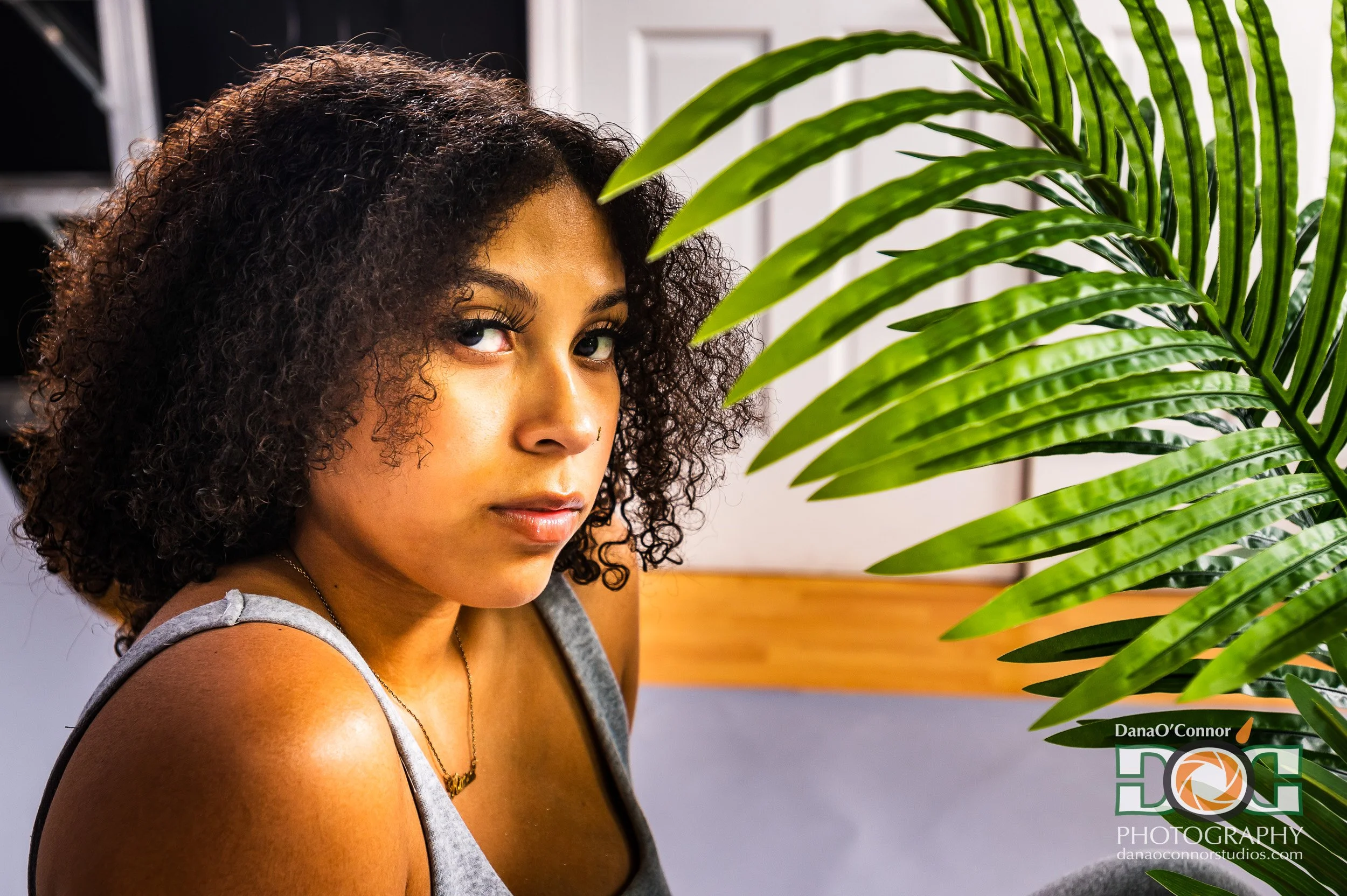 Portrait of a young woman with curly hair, gold hoop earrings, and a gray tank top looking towards a green tropical plant with long leaves in an indoor setting.