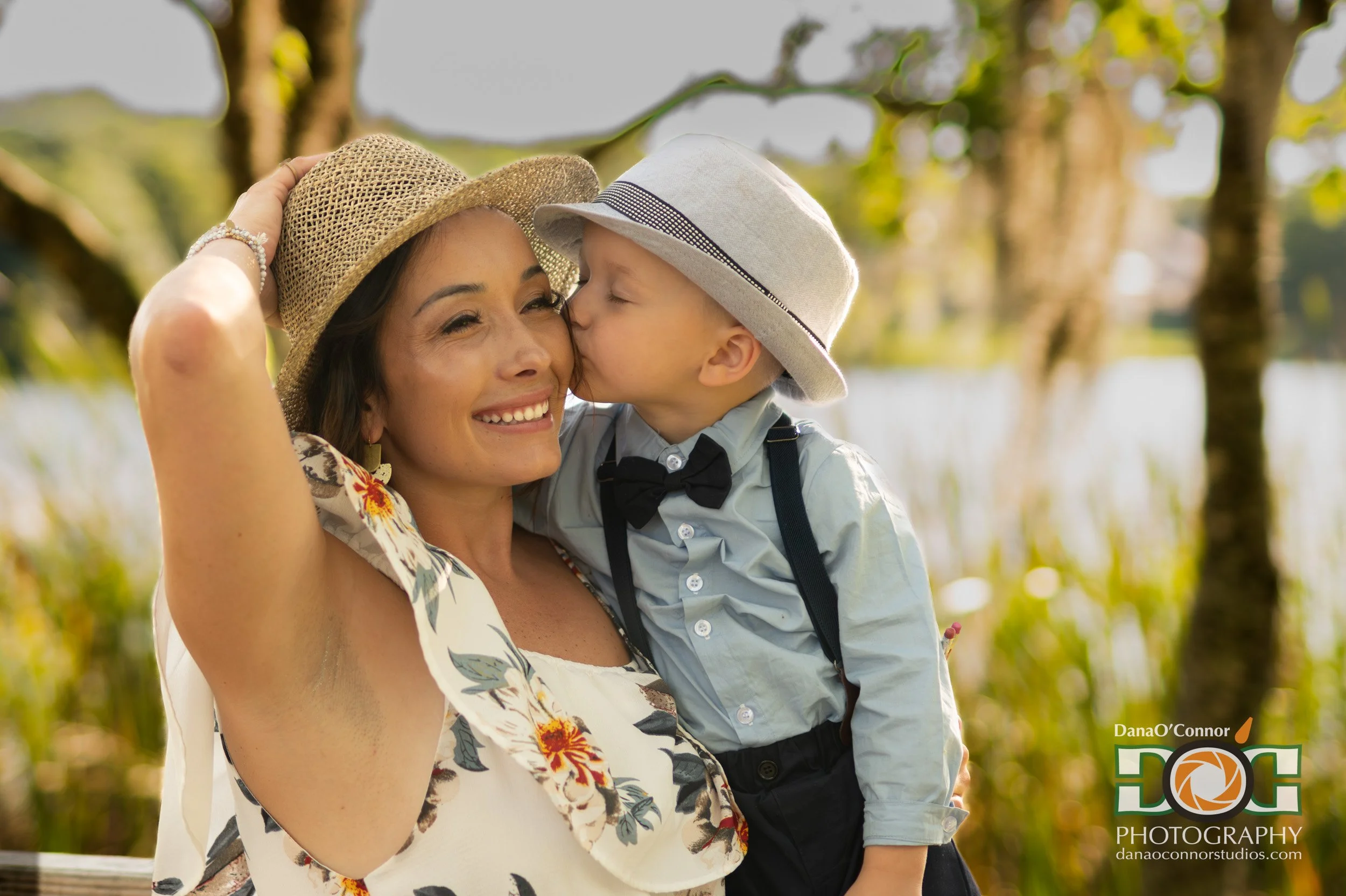 A woman smiling as a young boy kisses her on the cheek, both wearing hats outdoors with trees in the background.