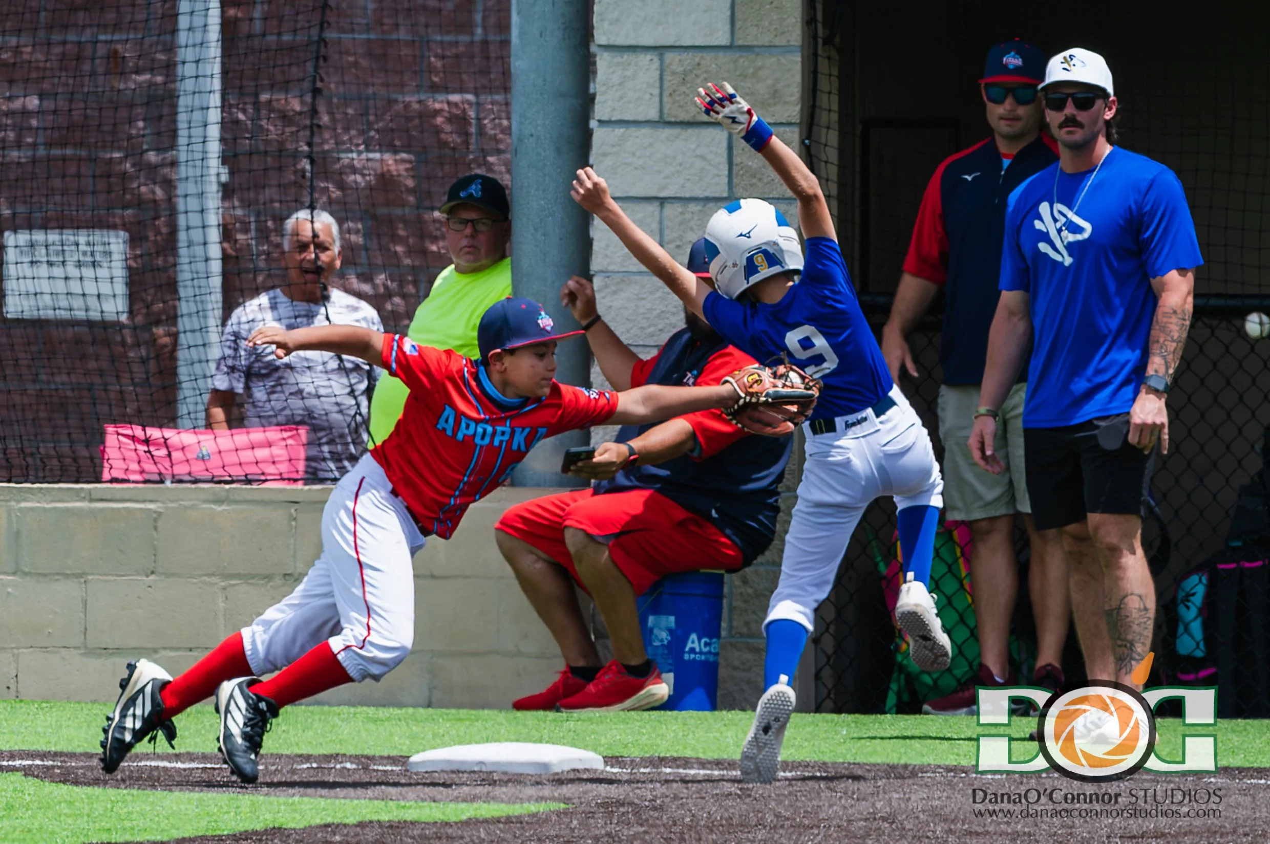 Apopka Titans 9U Travel Ball team's 3rd baseman, in a red and white uniform, dives to tag a baserunner,  in a blue uniform. Several onlookers, some wearing sunglasses, watch the play from behind a chain-link fence.