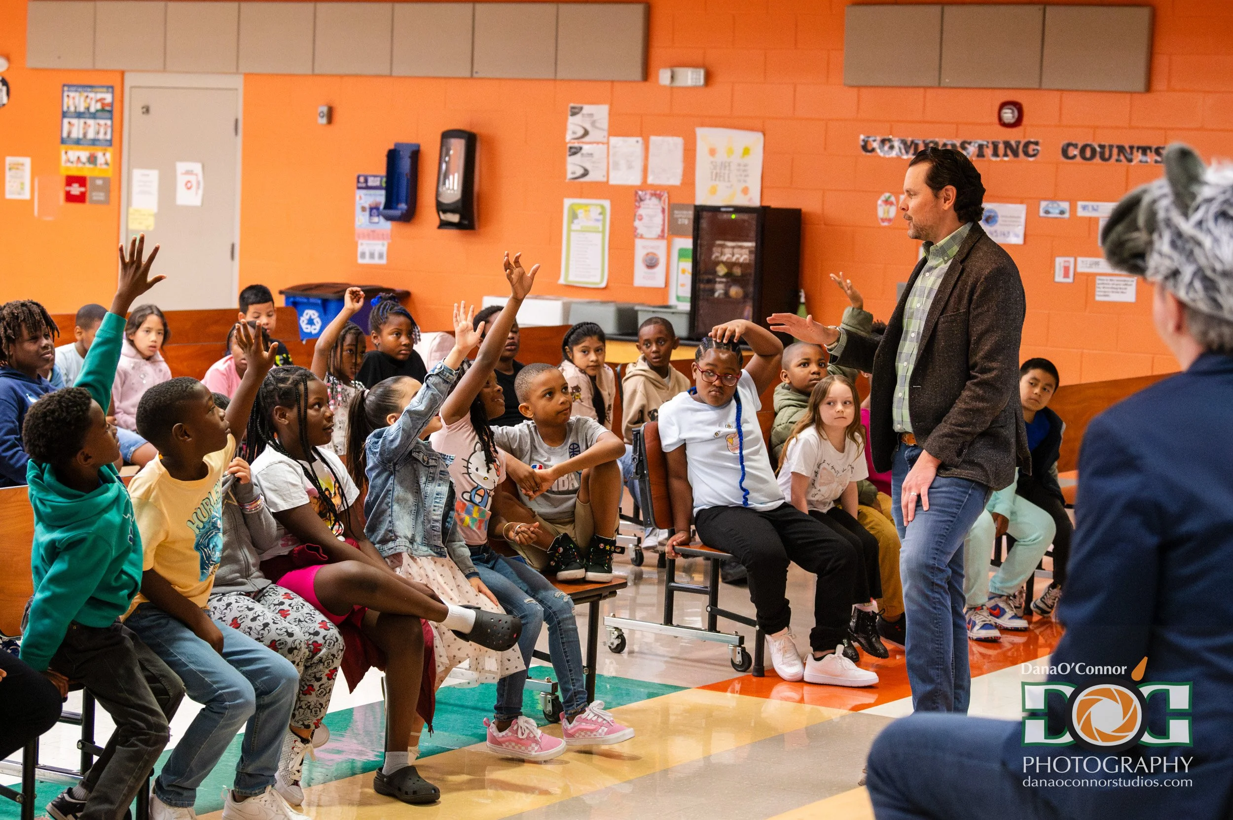 A man standing in front of a group of children sitting on chairs in a school gymnasium. The children are raising their hands, and the man appears to be engaging with them. The background shows orange walls with posters and a sign that says 'COMFORTIN