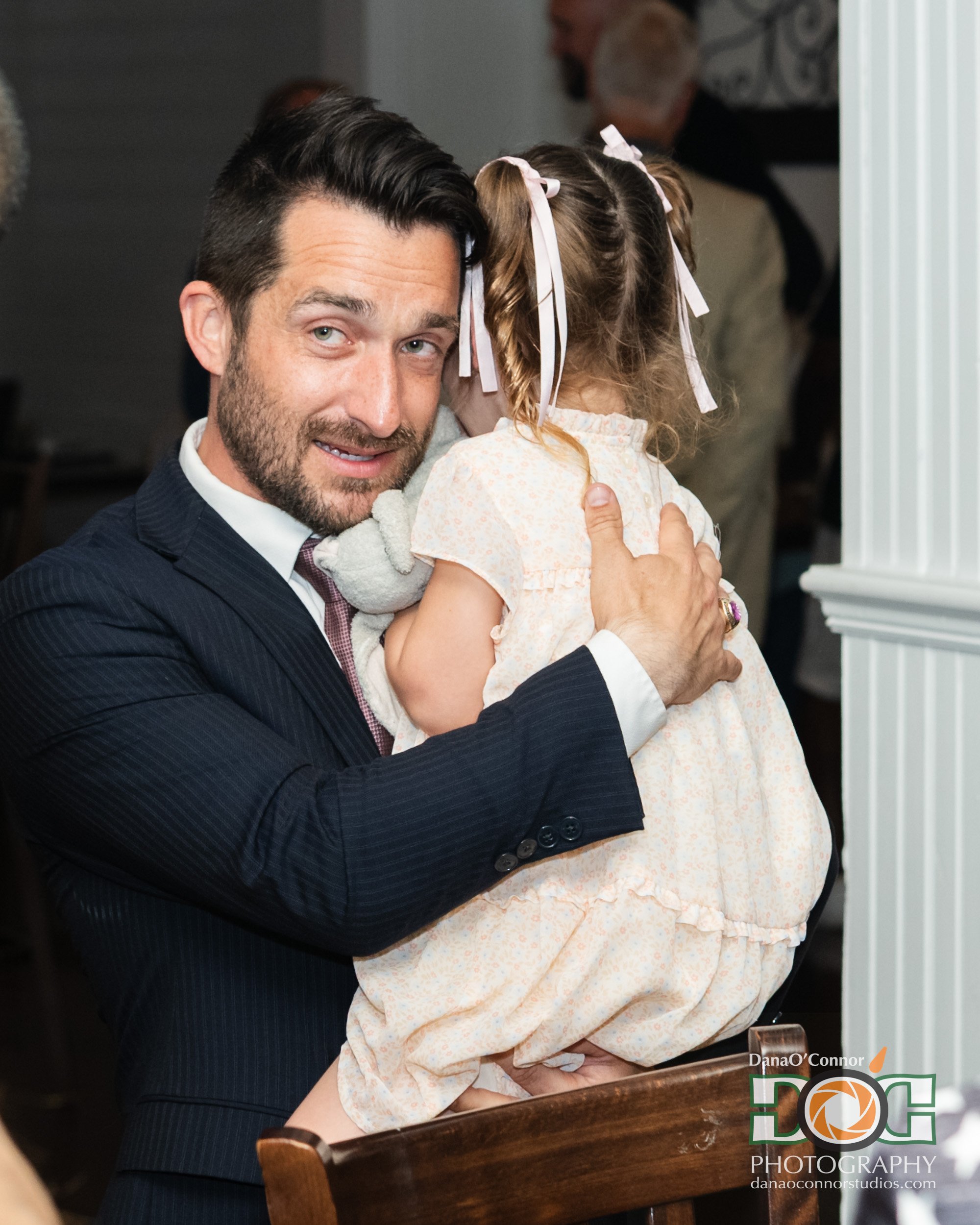 A man holding a young girl in his arms, indoors at a gathering or event. The girl has curly blonde hair tied with pink ribbons and is wearing a light-colored dress. The man is dressed in a dark suit and is smiling at the camera.