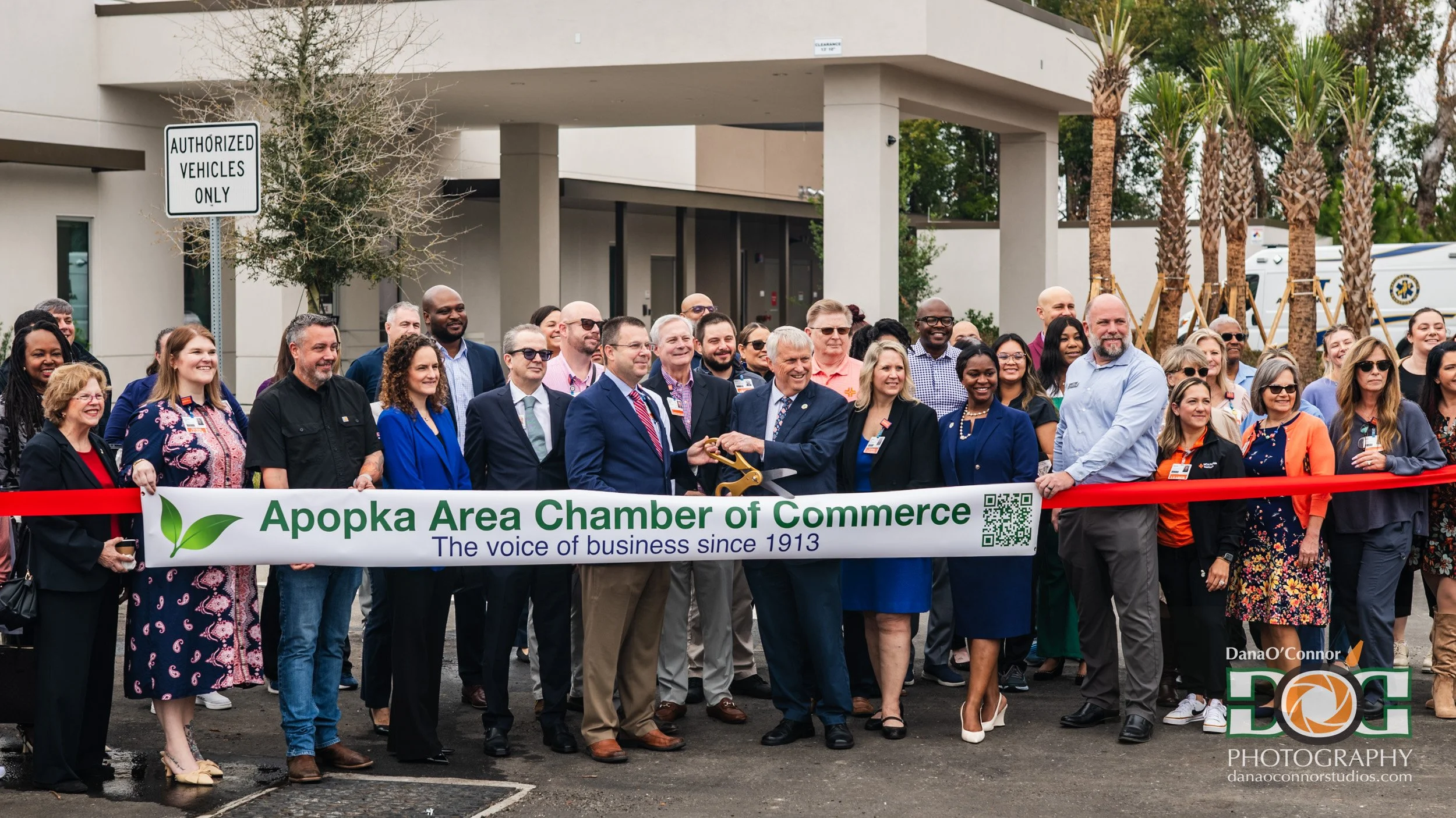Group of people at ribbon-cutting event holding a banner that reads 'Apopka Area Chamber of Commerce, The voice of business since 1913' in front of a modern building with palm trees.