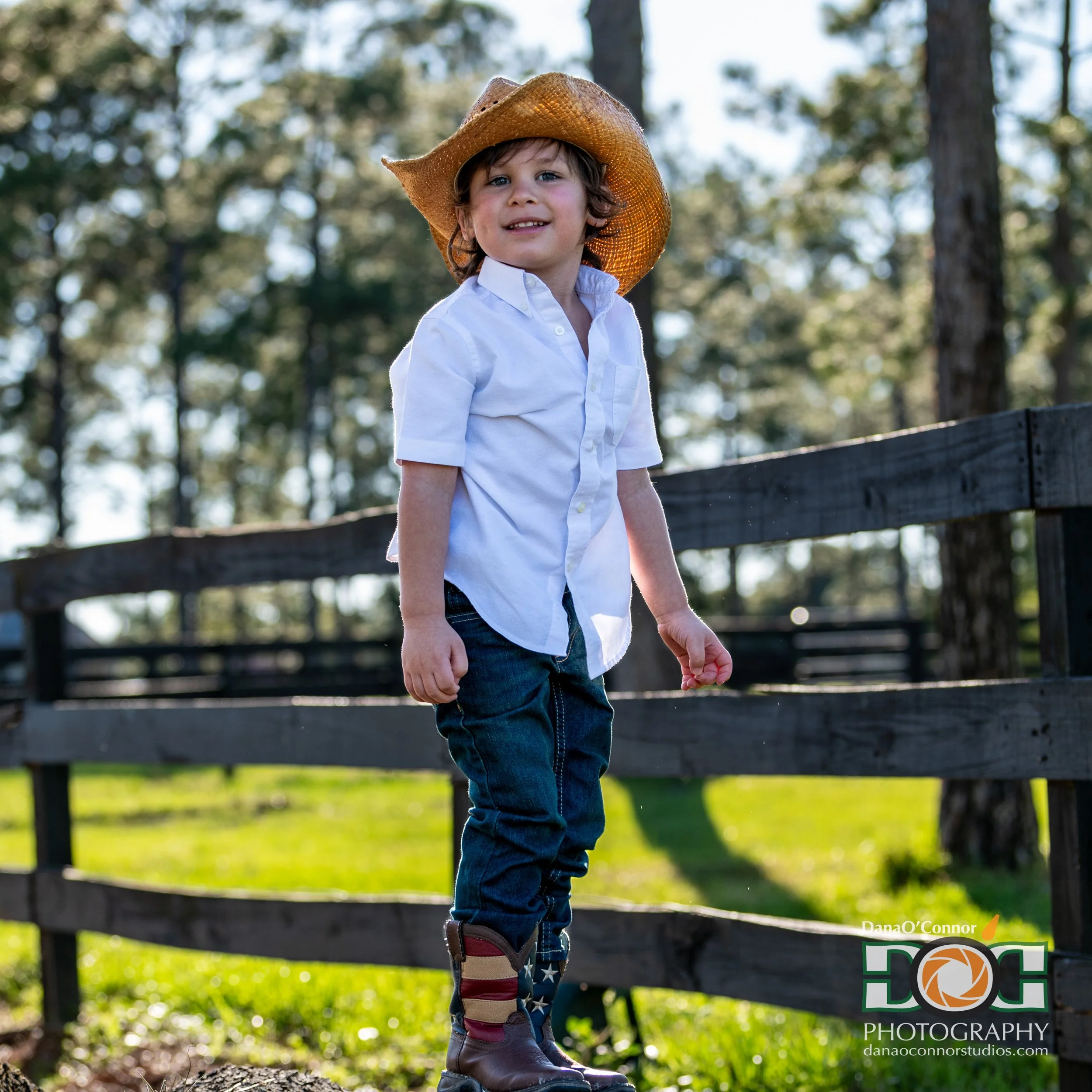 A young boy with curly hair wearing a white shirt, jeans, cowboy boots, and a straw hat, standing outdoors in front of a wooden fence and trees during daytime.