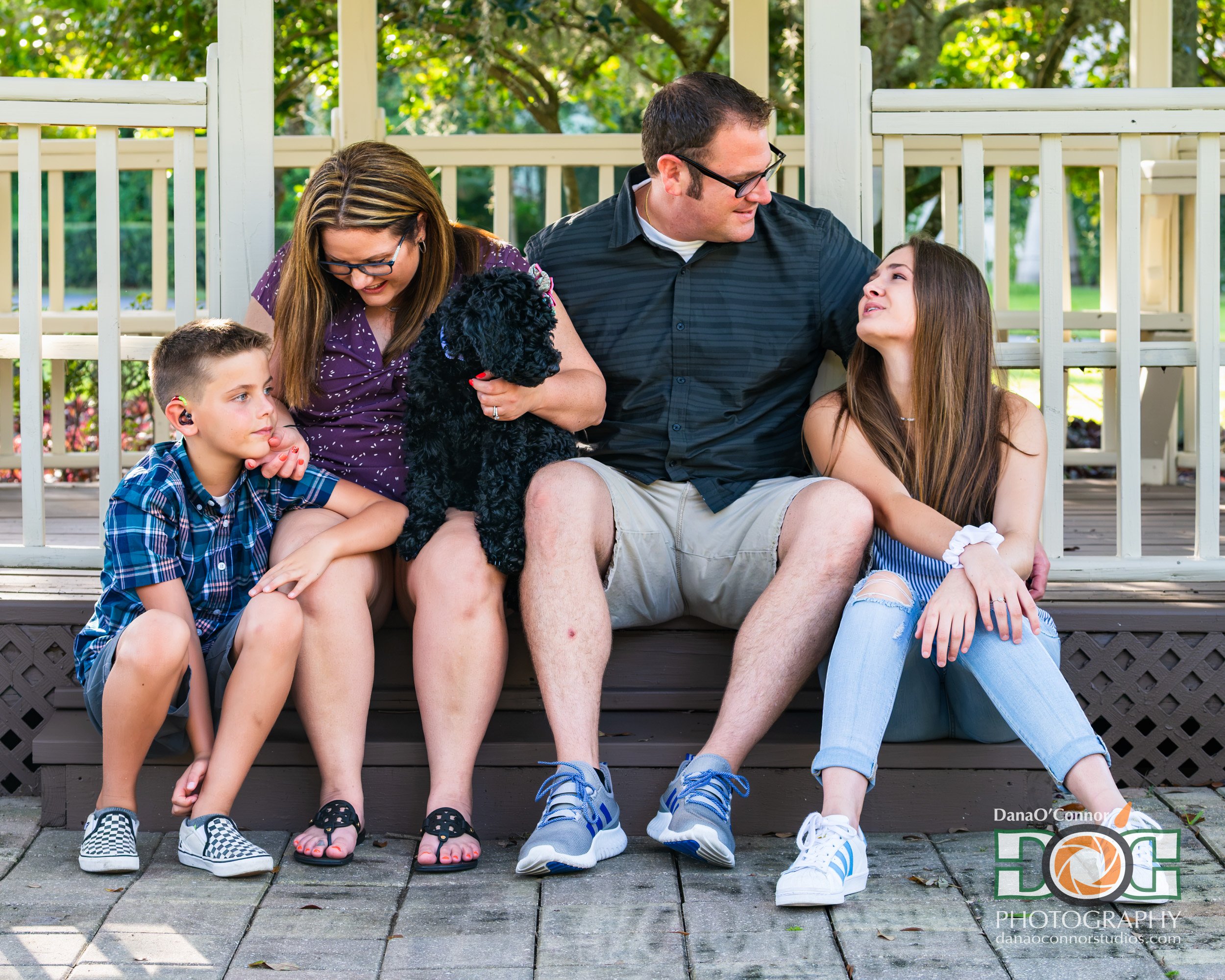 Family sitting on porch steps with two kids, a woman, a man, and a small black dog, outdoors with green trees in the background.
