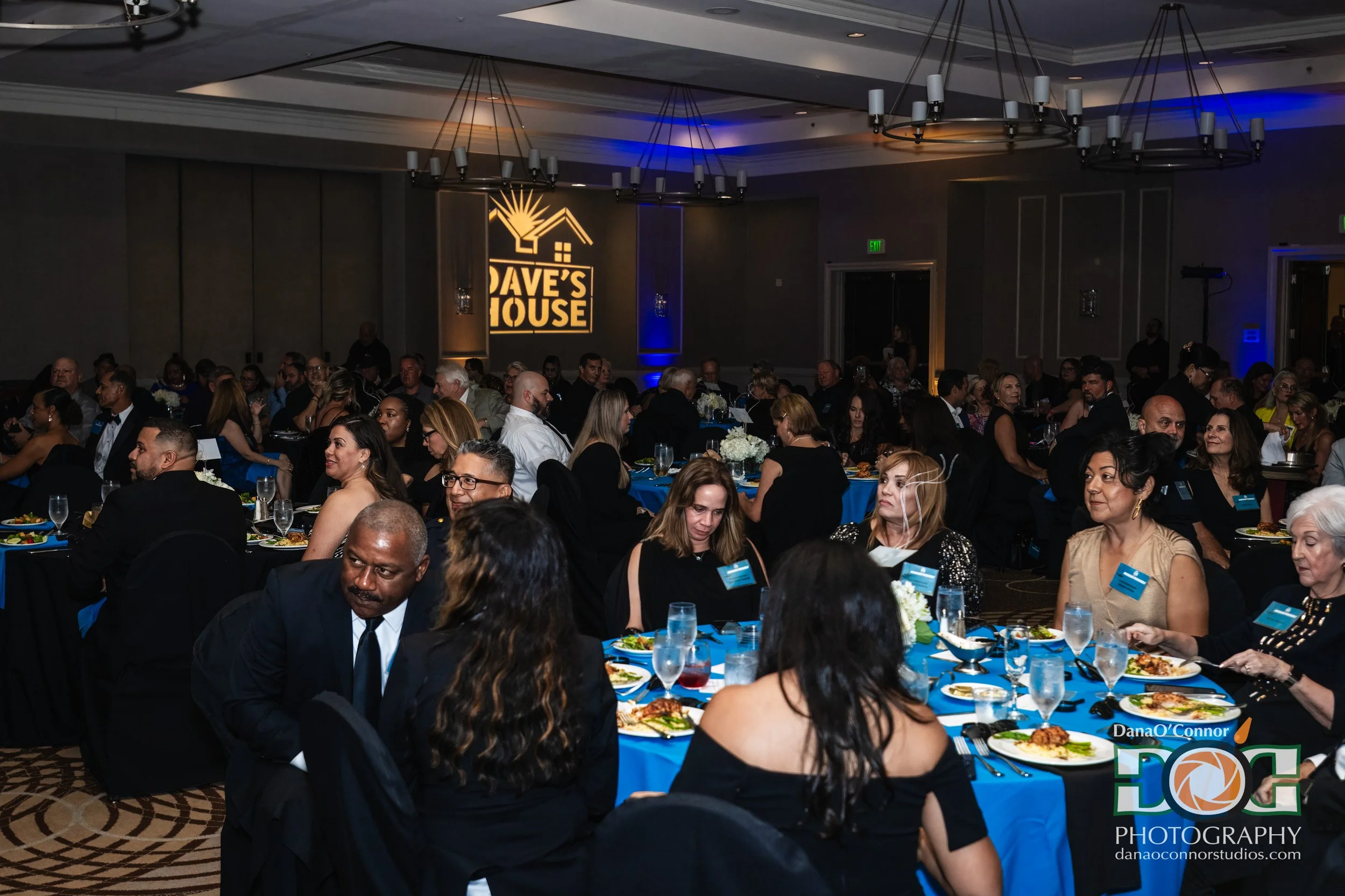 People attending a formal event or banquet seated at round tables with blue tablecloths, dining on plates of food. The room is decorated with chandeliers and uplighting, with a large sign reading 'Dave's House' on the wall. The scene is lively and we