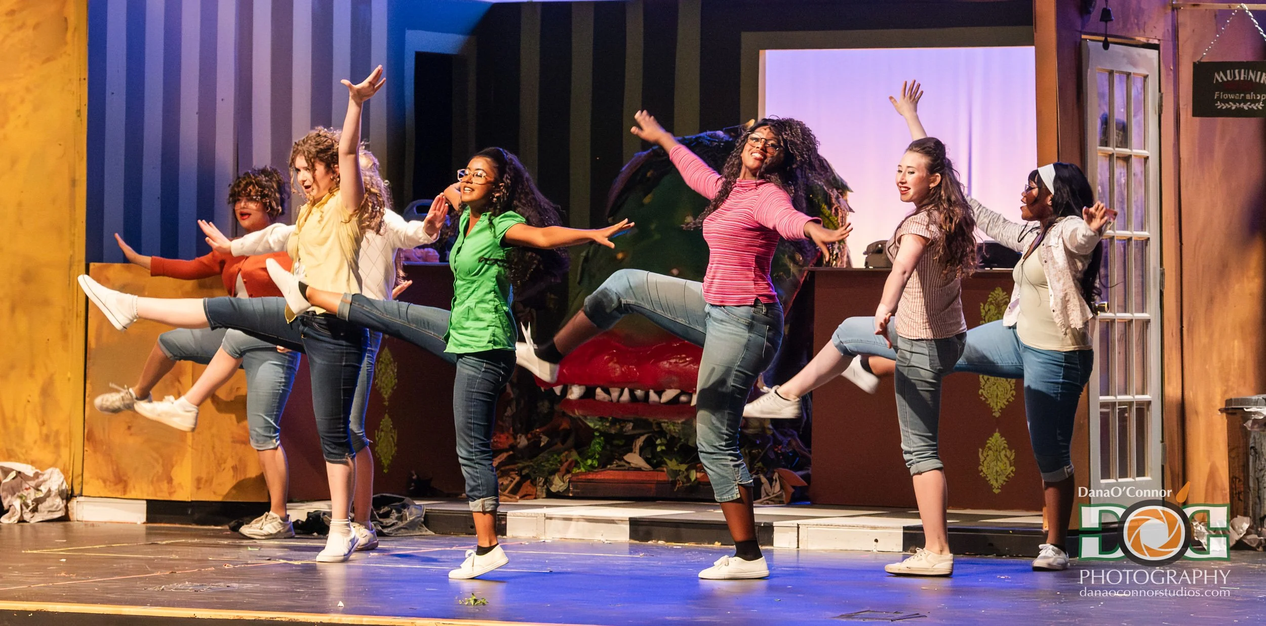 Group of women performing a dance on stage, with some lifting their legs and smiling, set against a colorful background with props that include a large red object and a door with a sign.