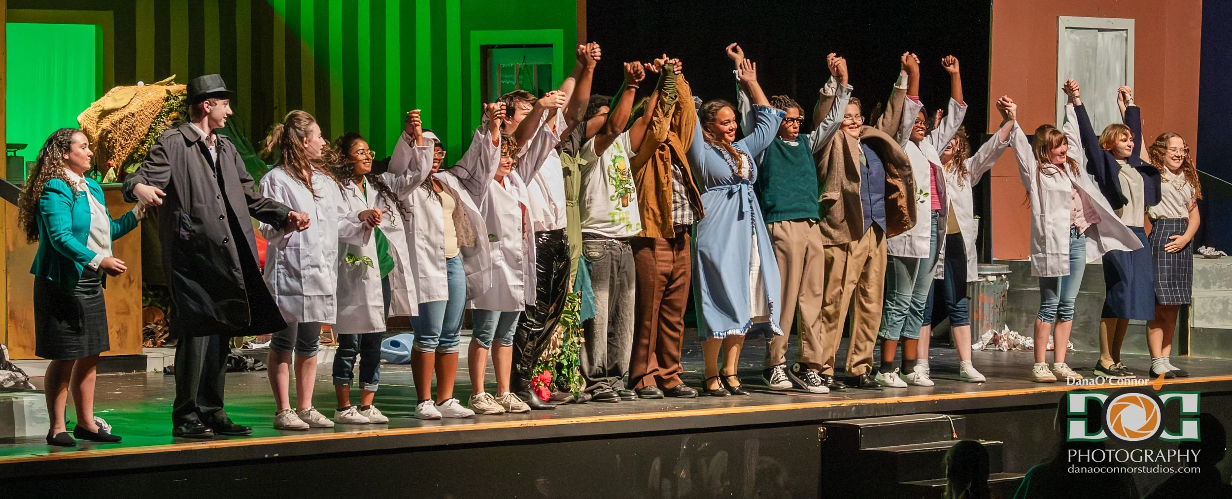 A diverse group of people on stage, holding hands and taking a bow after a performance, with colorful set design in the background.