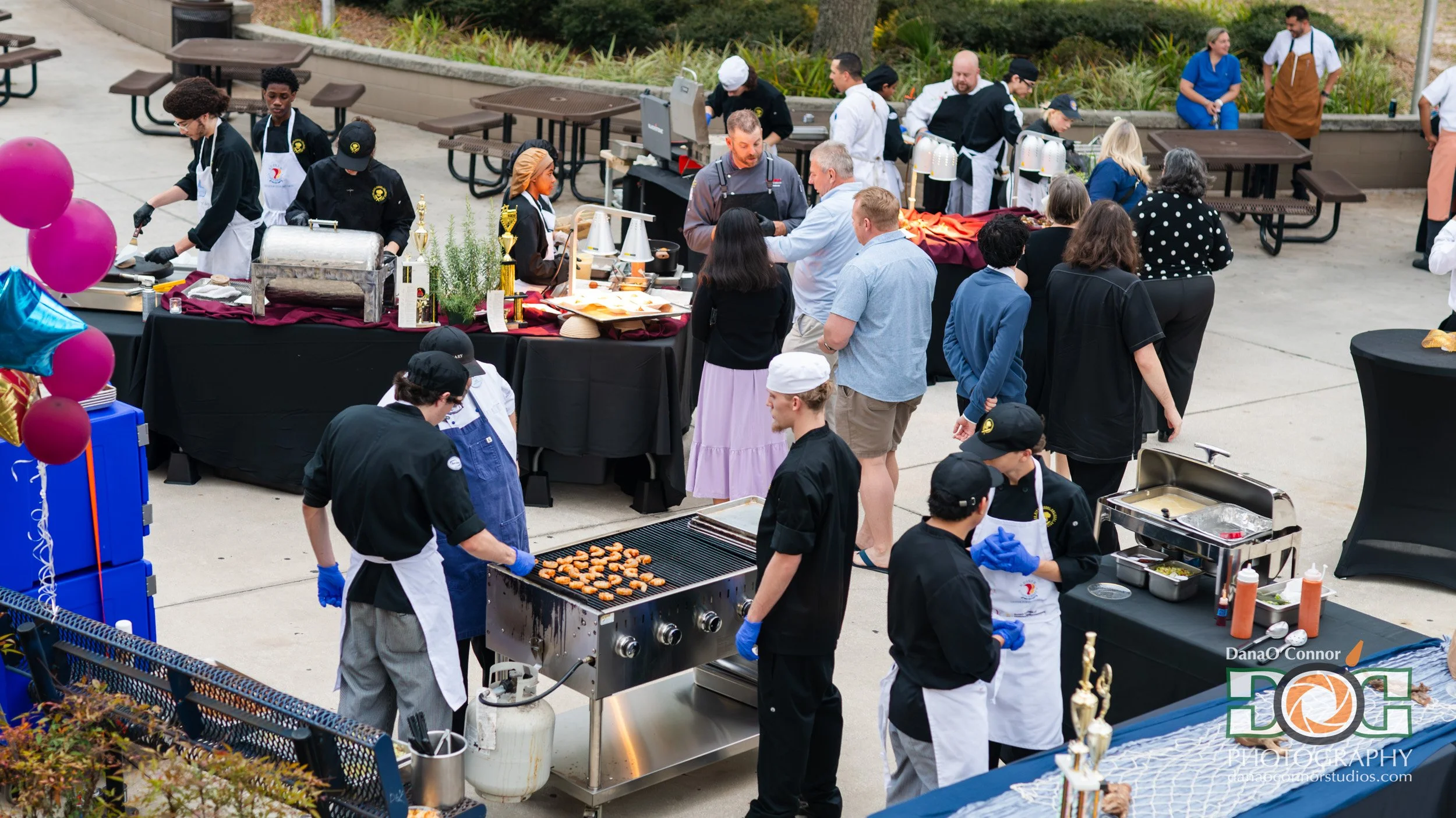A group of people including staff and guests at an outdoor buffet or food station during a celebration or event. Food is being grilled, and there are tables with dishes and trophies for awards.
