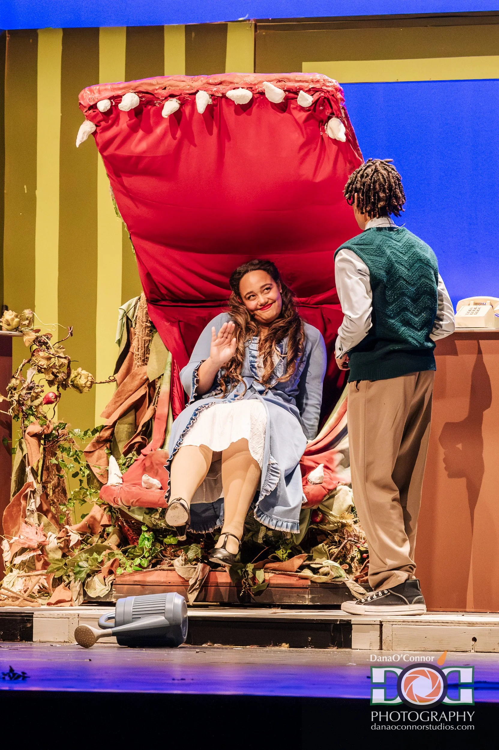 A theatrical scene featuring a woman sitting on a throne-like seat with a red canopy, signifying a dragon's mouth with white teeth. She is waving and smiling, dressed in period costume. A young man stands nearby in period attire, looking at her. The 