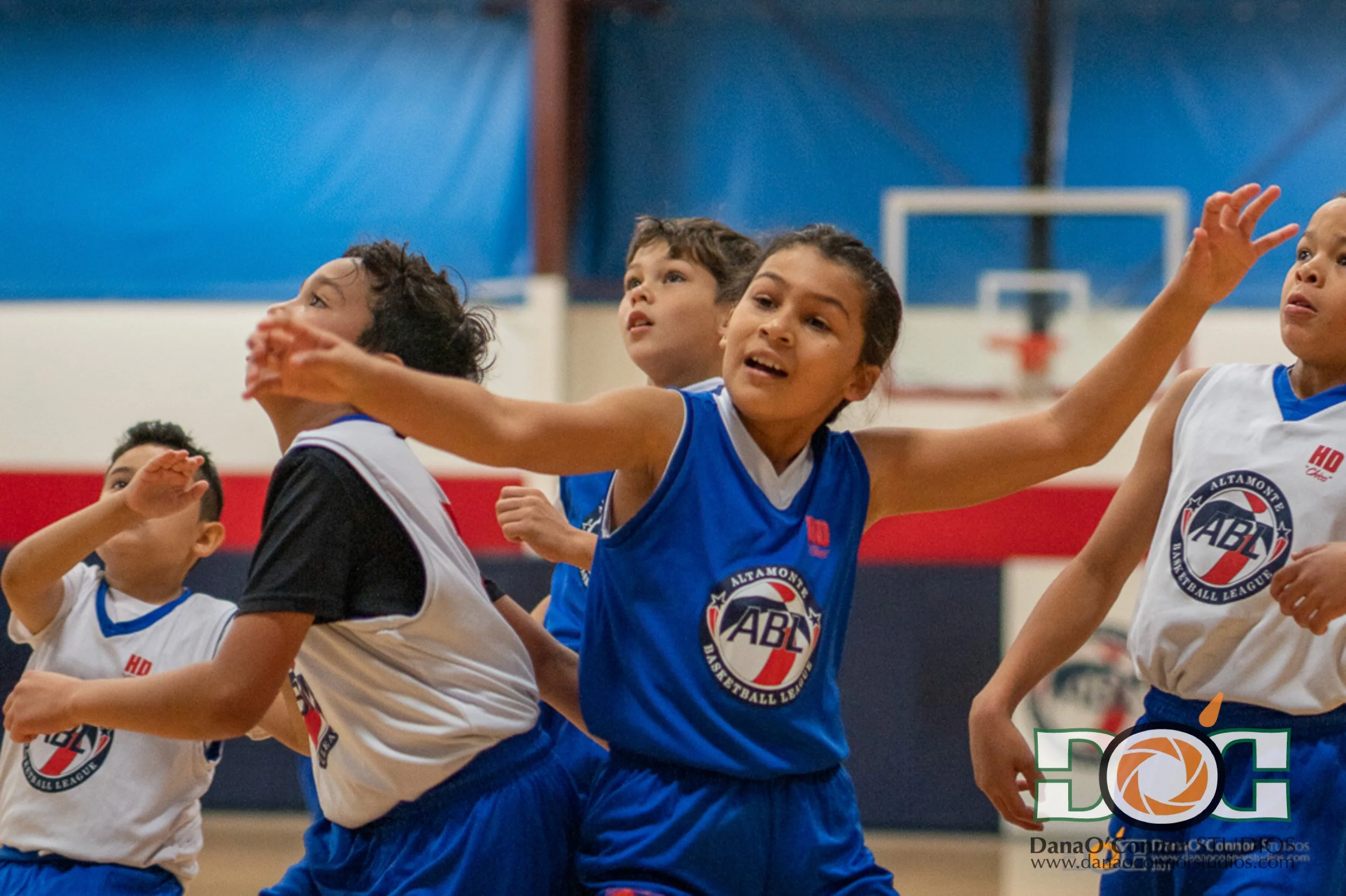 Children playing basketball during a game, wearing blue and white jerseys with the ABC Basketball League logo, in an indoor gymnasium.