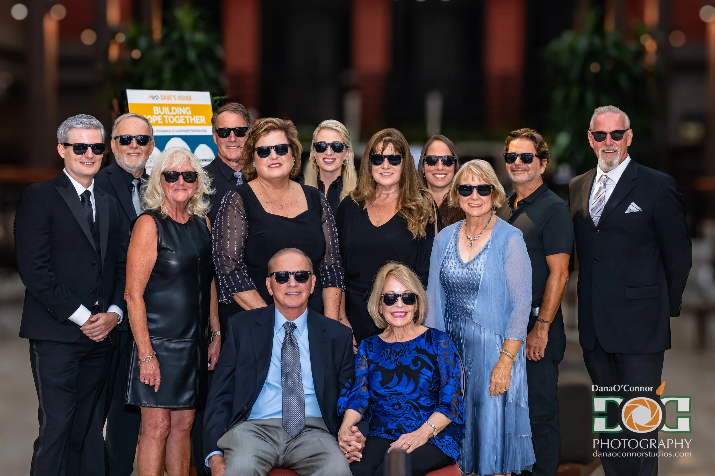 Group of people dressed in formal attire, wearing sunglasses, posing for a photo at an event, with a sign in the background that says 'Building Hope Together' and includes a logo. Multiple women and men of varying ages are smiling, some standing, som