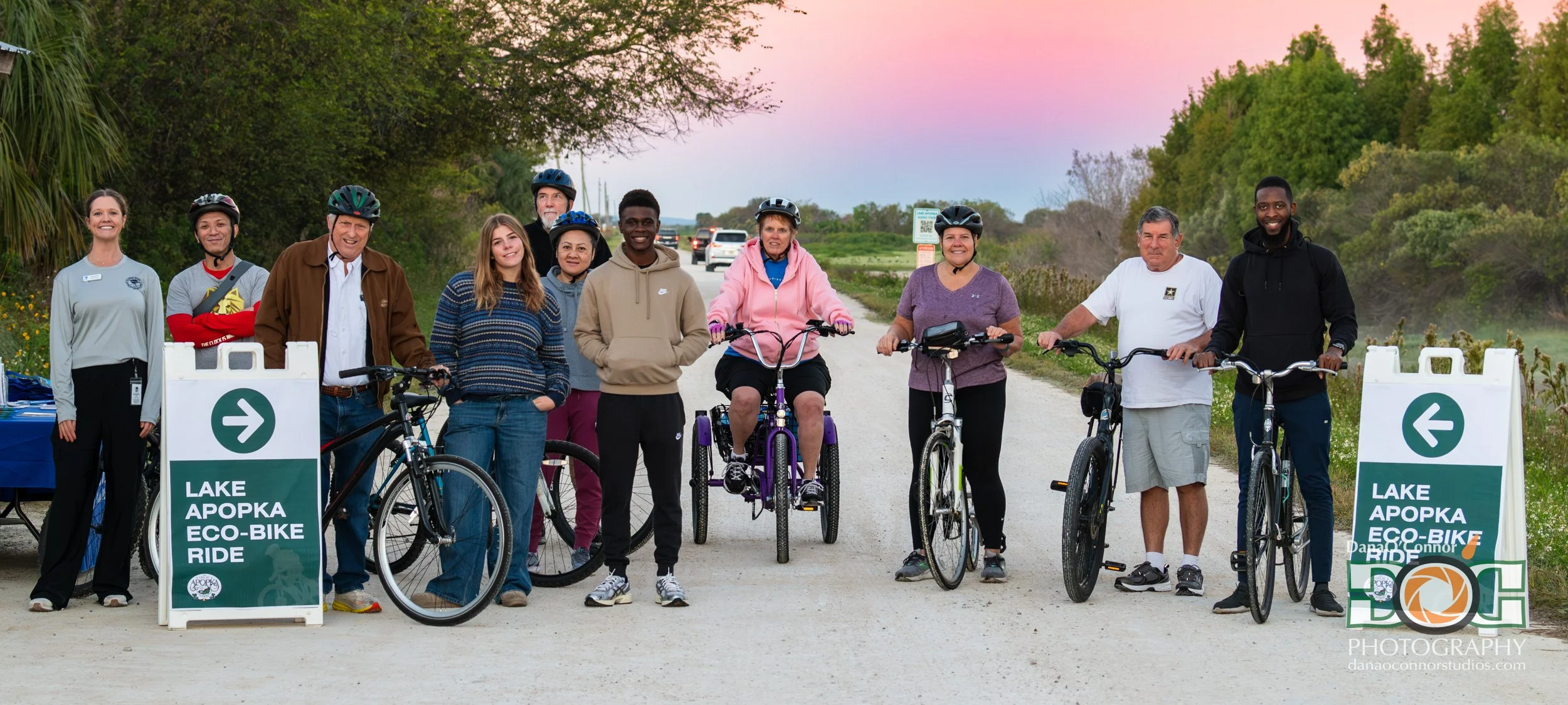 Group of people with bicycles participating in the Lake Apopka Eco-Bike Ride, standing on a dirt path with lush greenery and a pink-tinged sky in the background.
