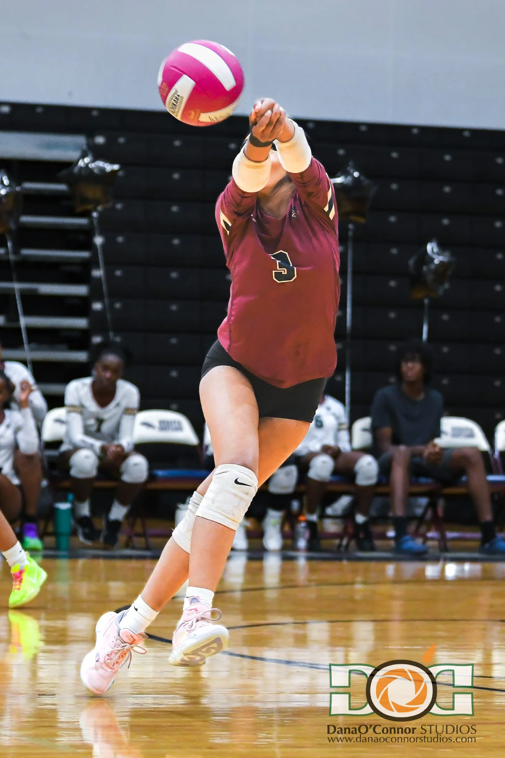 A Wekiva High School female volleyball player in a maroon jersey with the number 5, hitting a pink and white volleyball during a game, with her teammates sitting on the bench in the background.