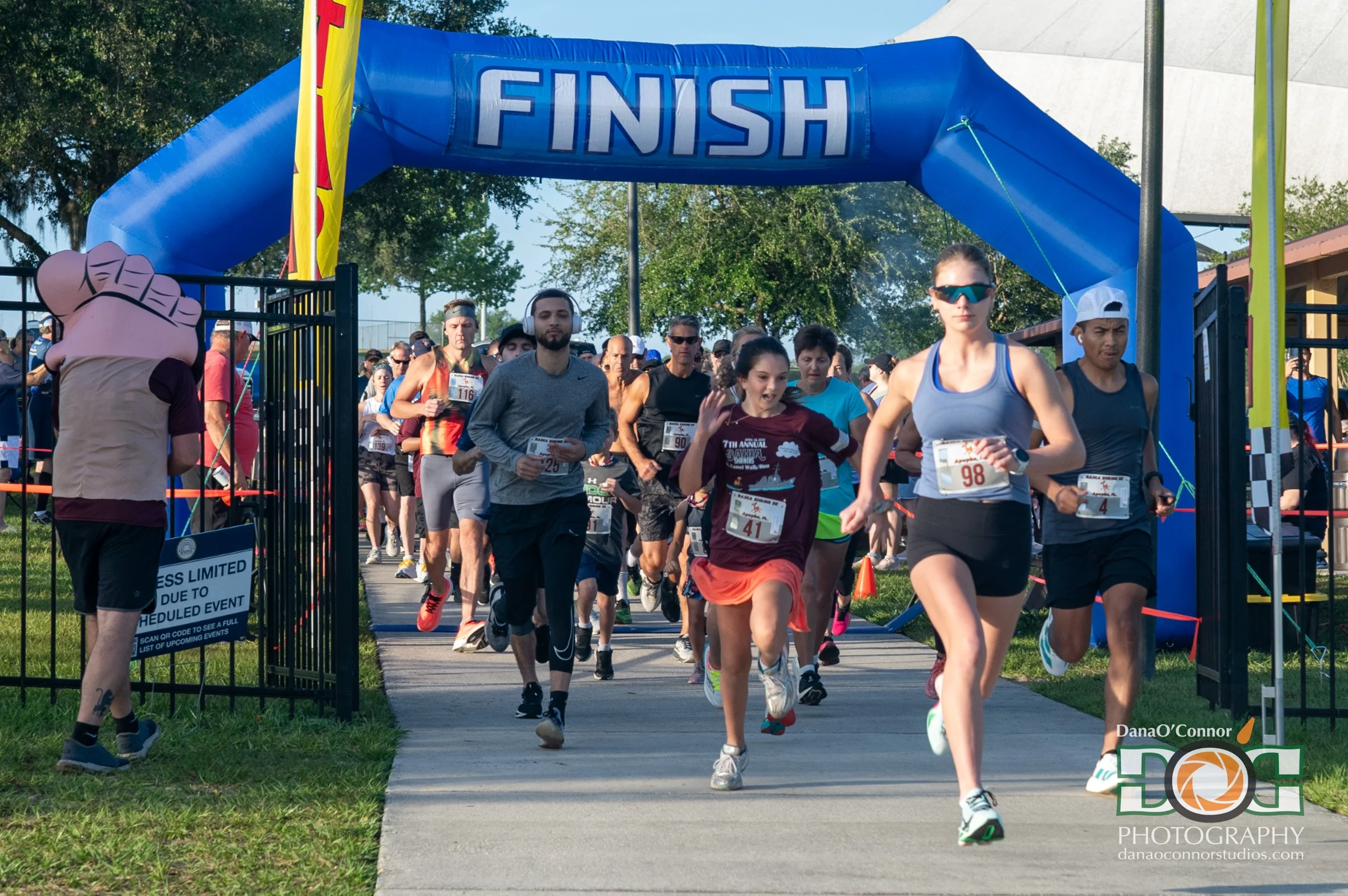 A crowd of runners exits a blue inflatable arch with a "FINISH" sign; some wear race bibs, and a mascot figure is visible near the gate.