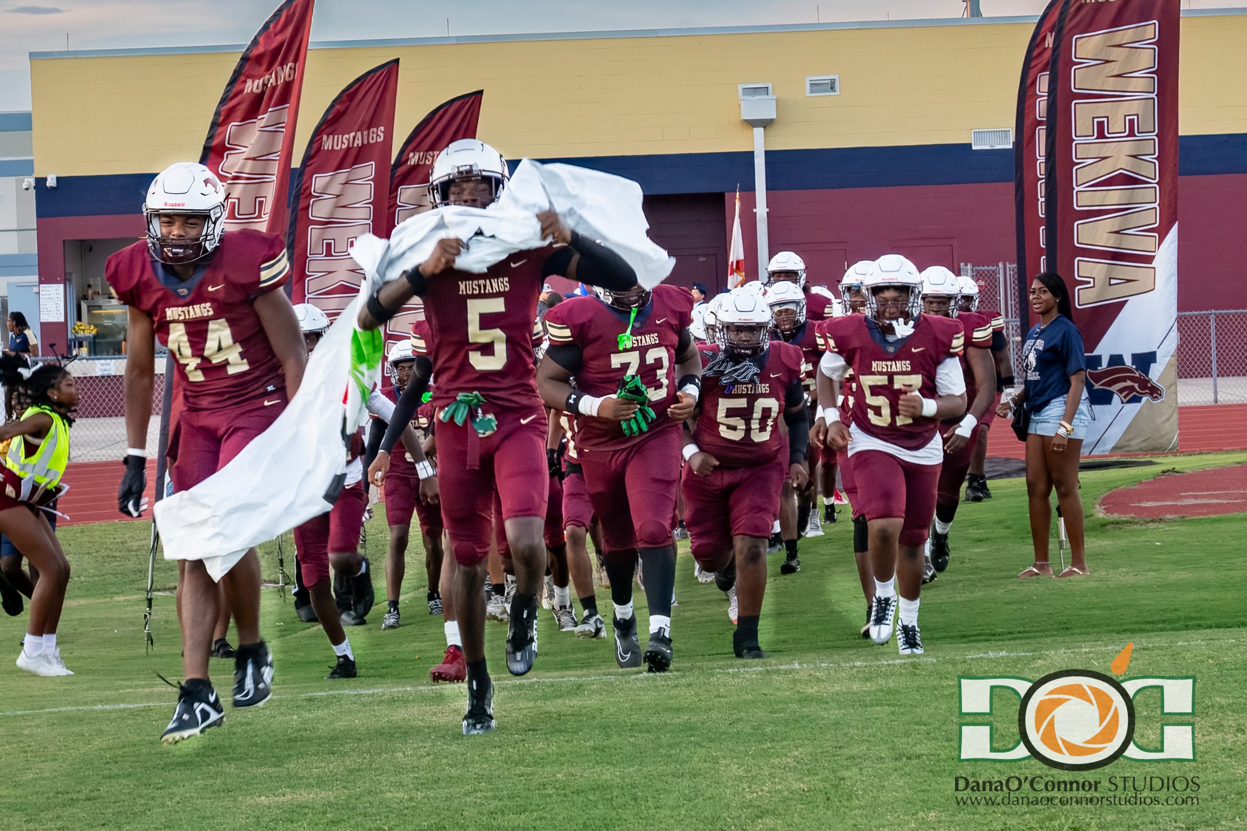 Wekiva High School football team in maroon uniforms running onto the field through a tunnel of banners with 'Mustangs' written on them. A woman and a young girl are on the sidelines.