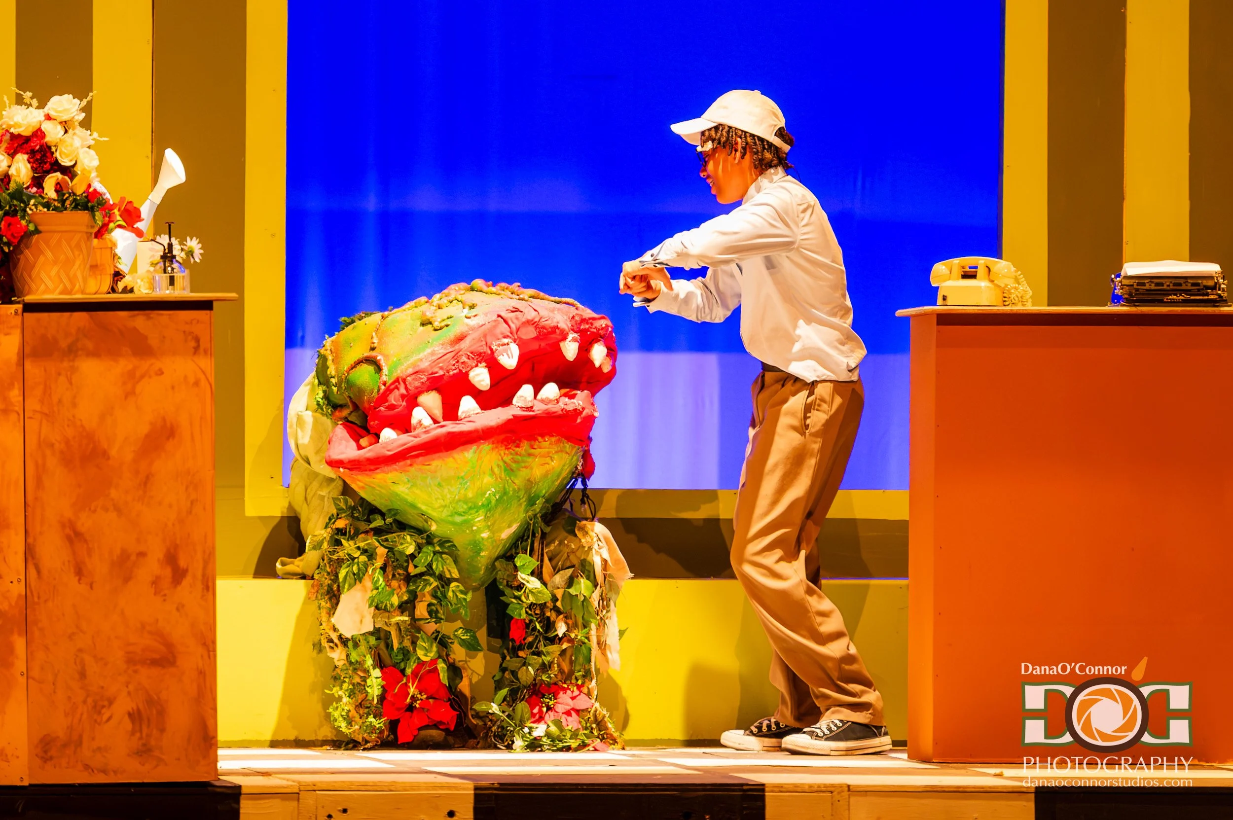 A person wearing a white cap and beige pants, with glasses and dreadlocks, is working on a colorful, large, dragon-like puppet on stage with a blue backdrop. There are flowers on the left and a yellow wall in the background.