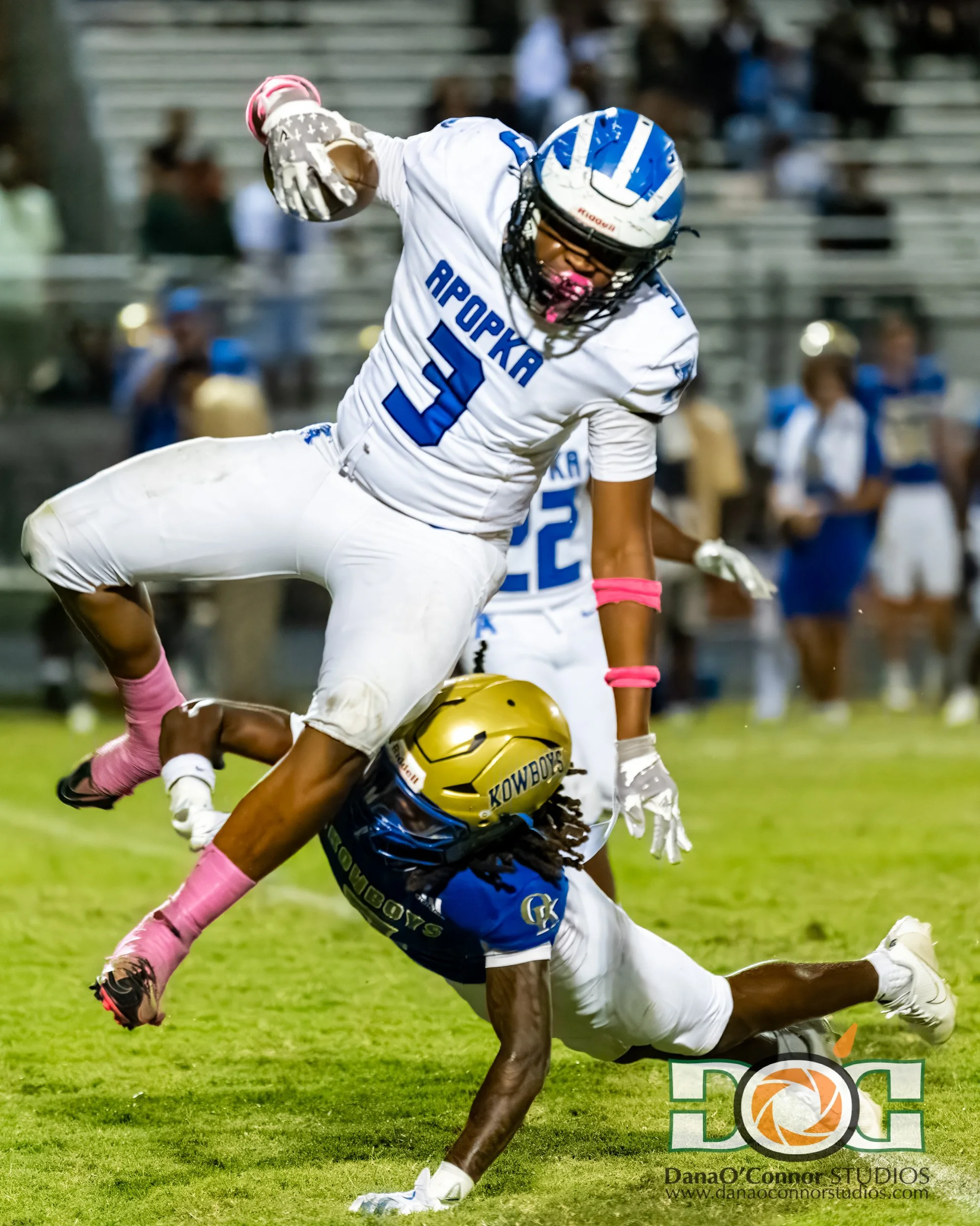 Two football players from opposing teams are colliding during a game on a grassy field at night. One player, wearing a blue and white uniform with a gold helmet labeled 'KOWBOYS,' is falling to the ground. The other player, in a white uniform with bl