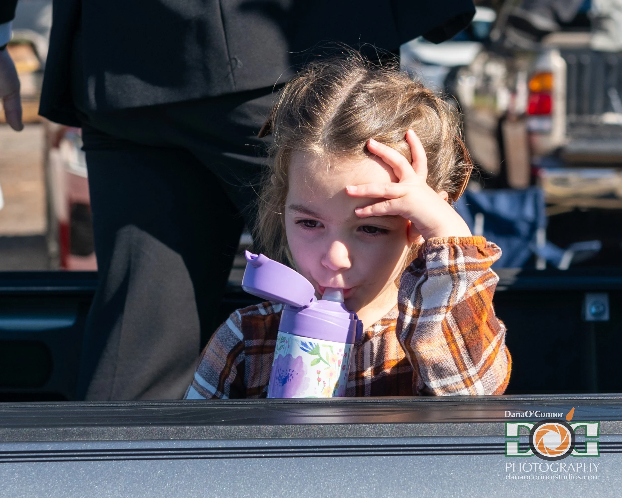 A young girl with light brown hair and light skin standing inside a vehicle, holding her forehead with one hand and drinking from a pink and floral cup, wearing a brown plaid shirt. The vehicle's door frame is visible at the bottom of the image.