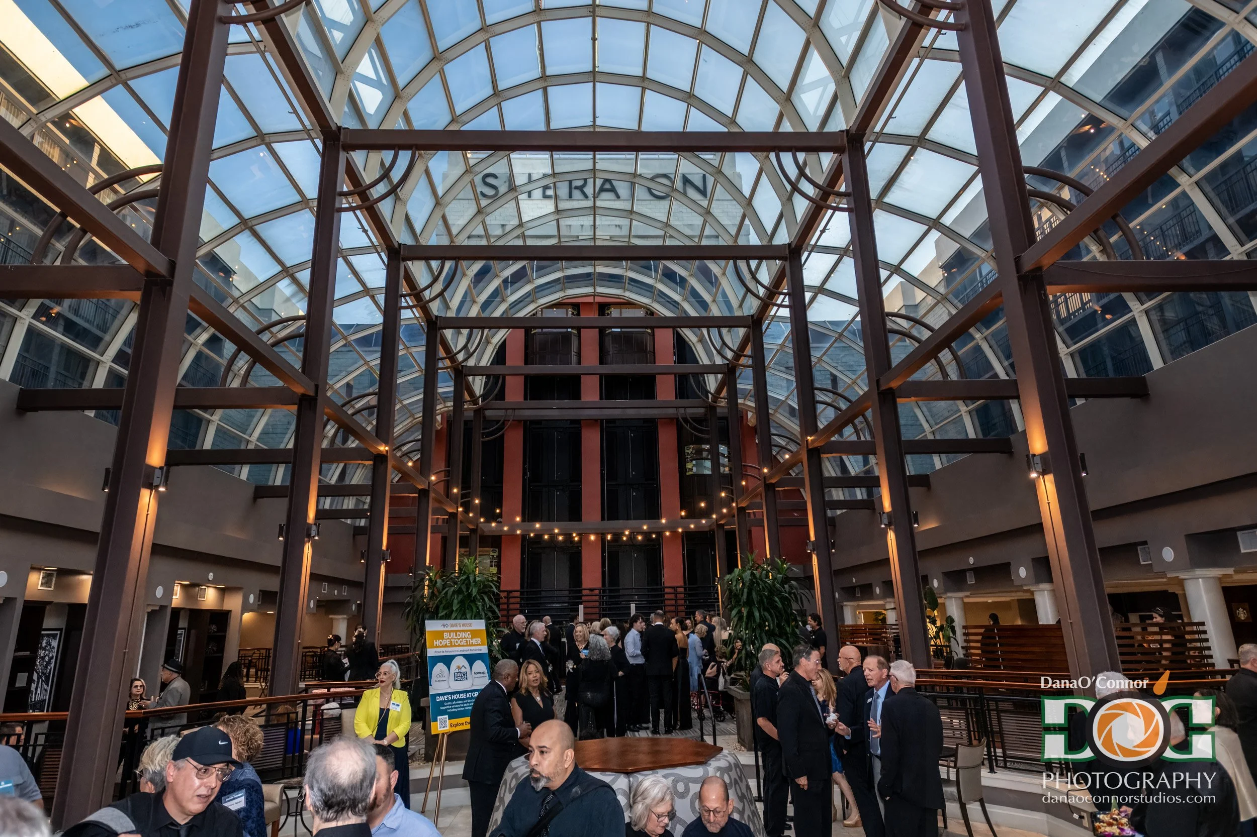 People gather inside a large, glass-covered atrium with a high arched ceiling, modern architectural design, and some standing near informational signboards.
