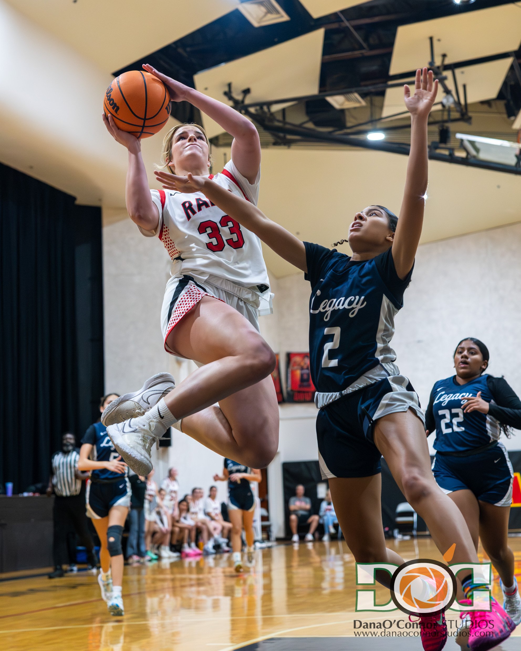 A female basketball player in a white and red uniform, number 33, jumping to shoot the ball on an indoor basketball court. She is surrounded by other players, including a girl in a navy uniform attempting to block her shot, and another girl with jers