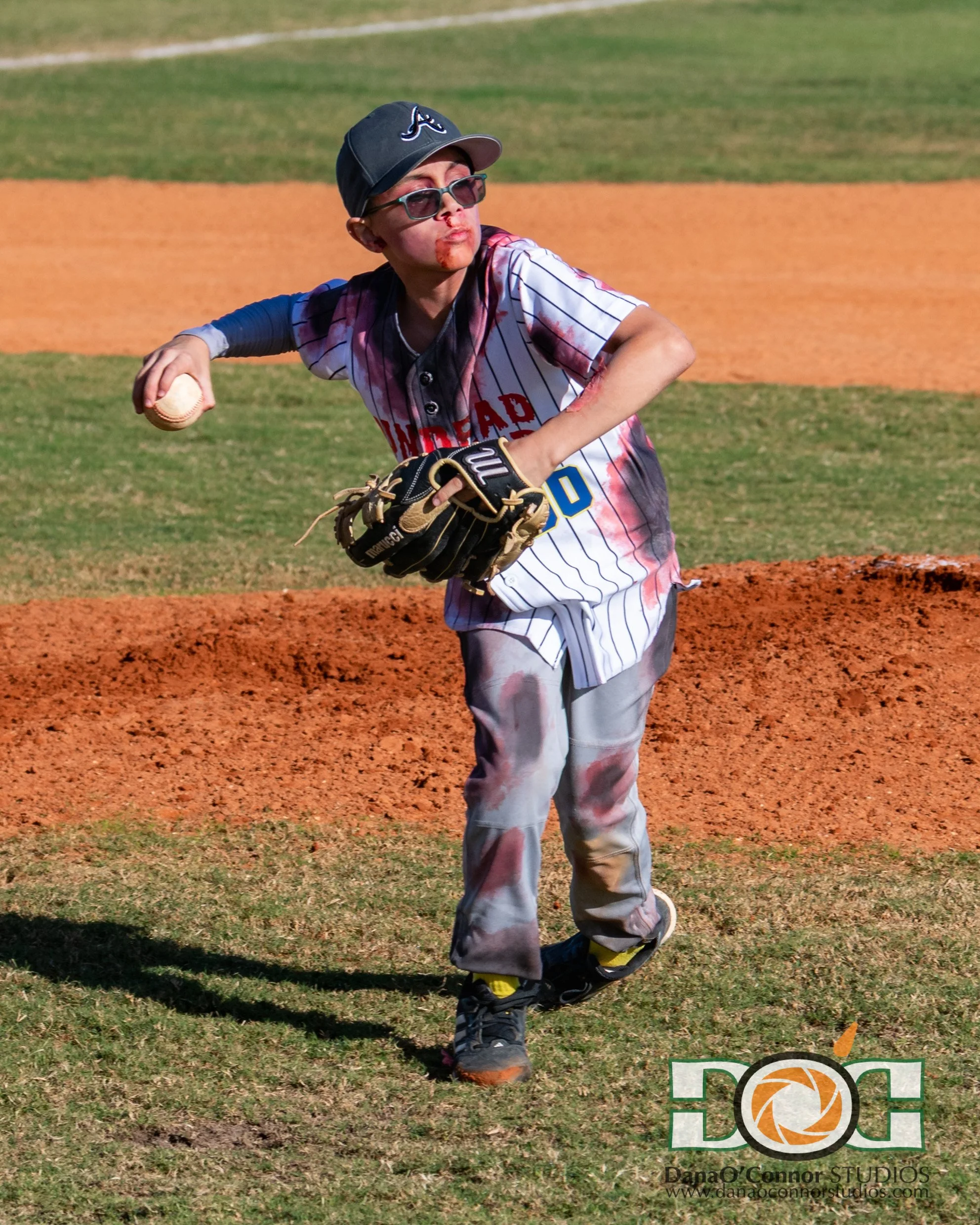 Young boy playing baseball on a field, wearing sunglasses, a baseball cap, and a baseball uniform with bloodstains, holding a baseball in his right hand, standing in a batting stance with a baseball glove on his left hand, on grass with a dirt infiel