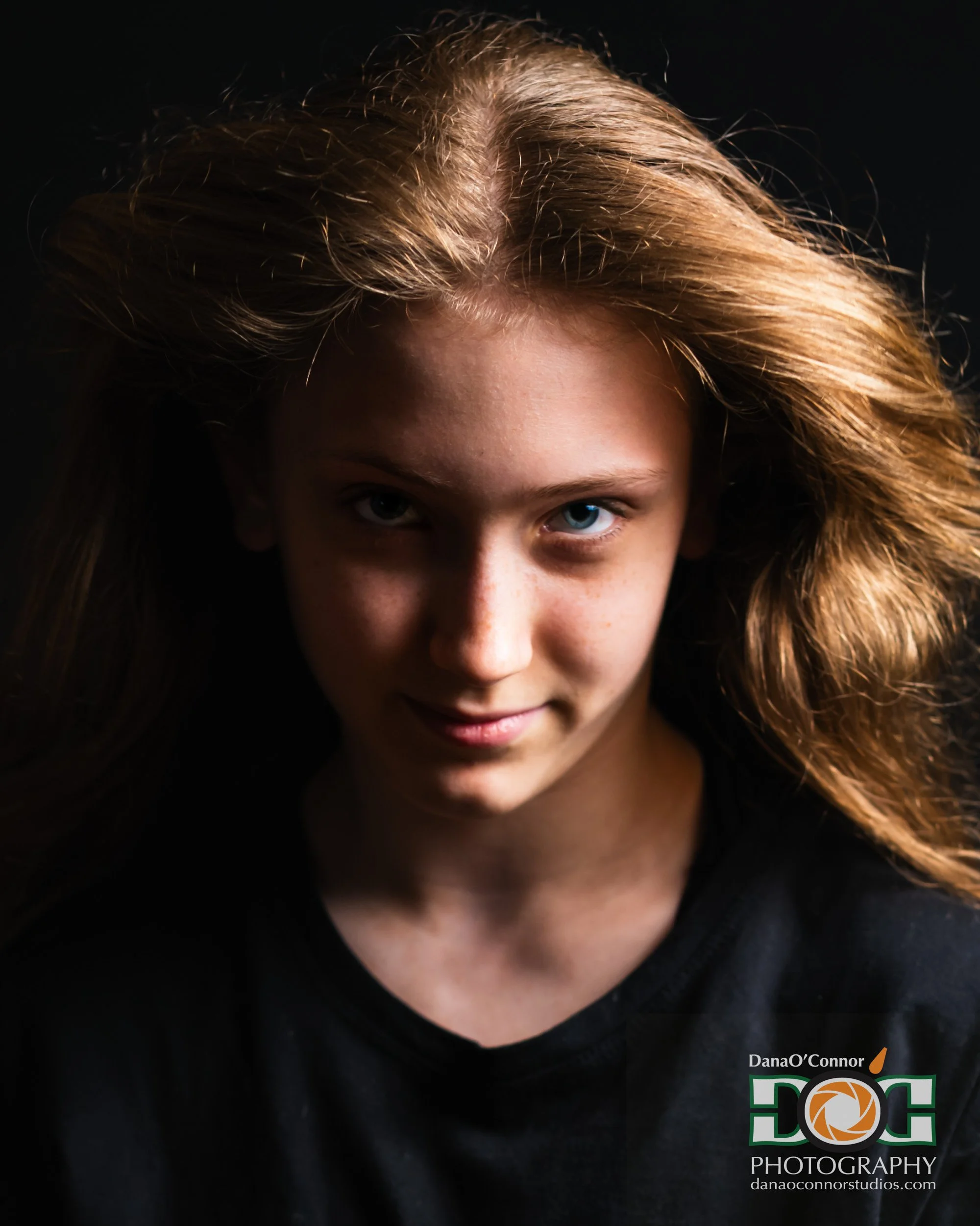 Portrait of a young woman with long, light brown hair and blue eyes, wearing a black shirt with a logo for a photography studio.