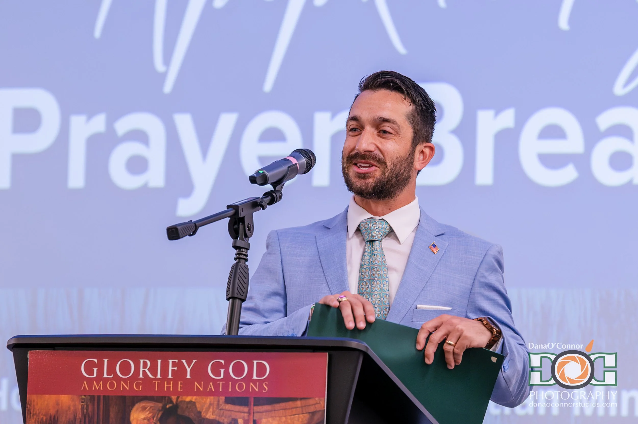 A man with dark hair and a beard wearing a light blue suit and a patterned tie speaking at a podium with a microphone. Behind him is a large screen displaying the words 'Traveling' and part of a phrase 'Glorify God among the nations'. The podium has 