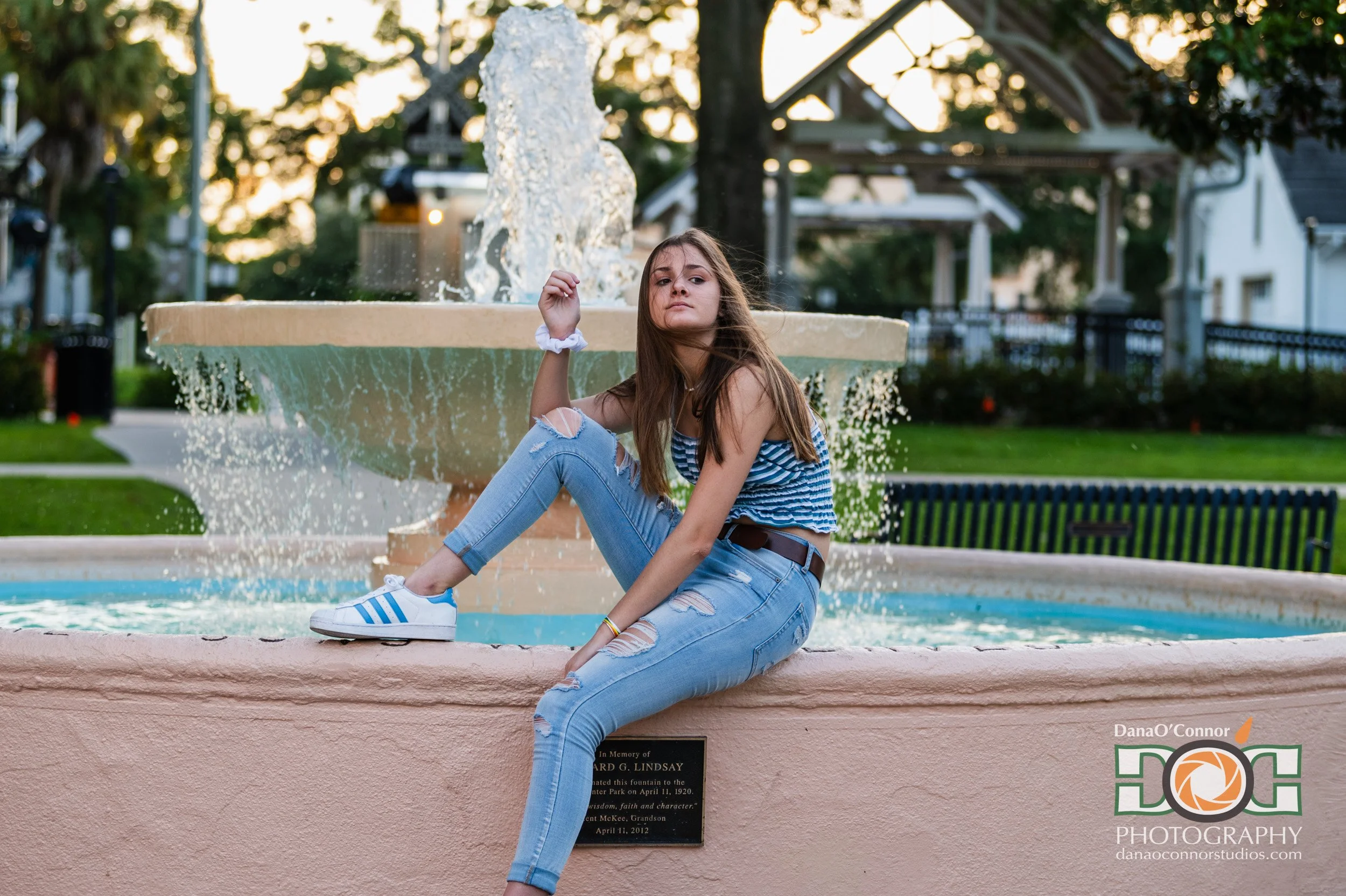 A young woman with long brown hair, wearing a striped top and ripped jeans, sitting on a pink fountain ledge in a park at sunset, with water splashing from the fountain behind her.