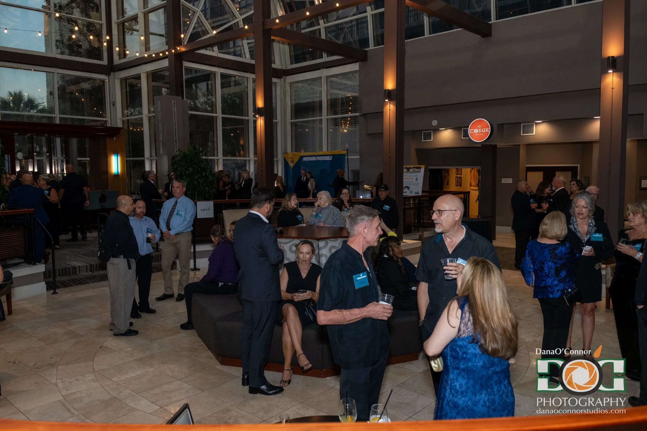 A group of people gathered in a large indoor space with high glass walls and ceiling, decorated with string lights. Some people are standing, others are sitting and chatting, holding drinks. There is a banner in the background with the words 'Mission