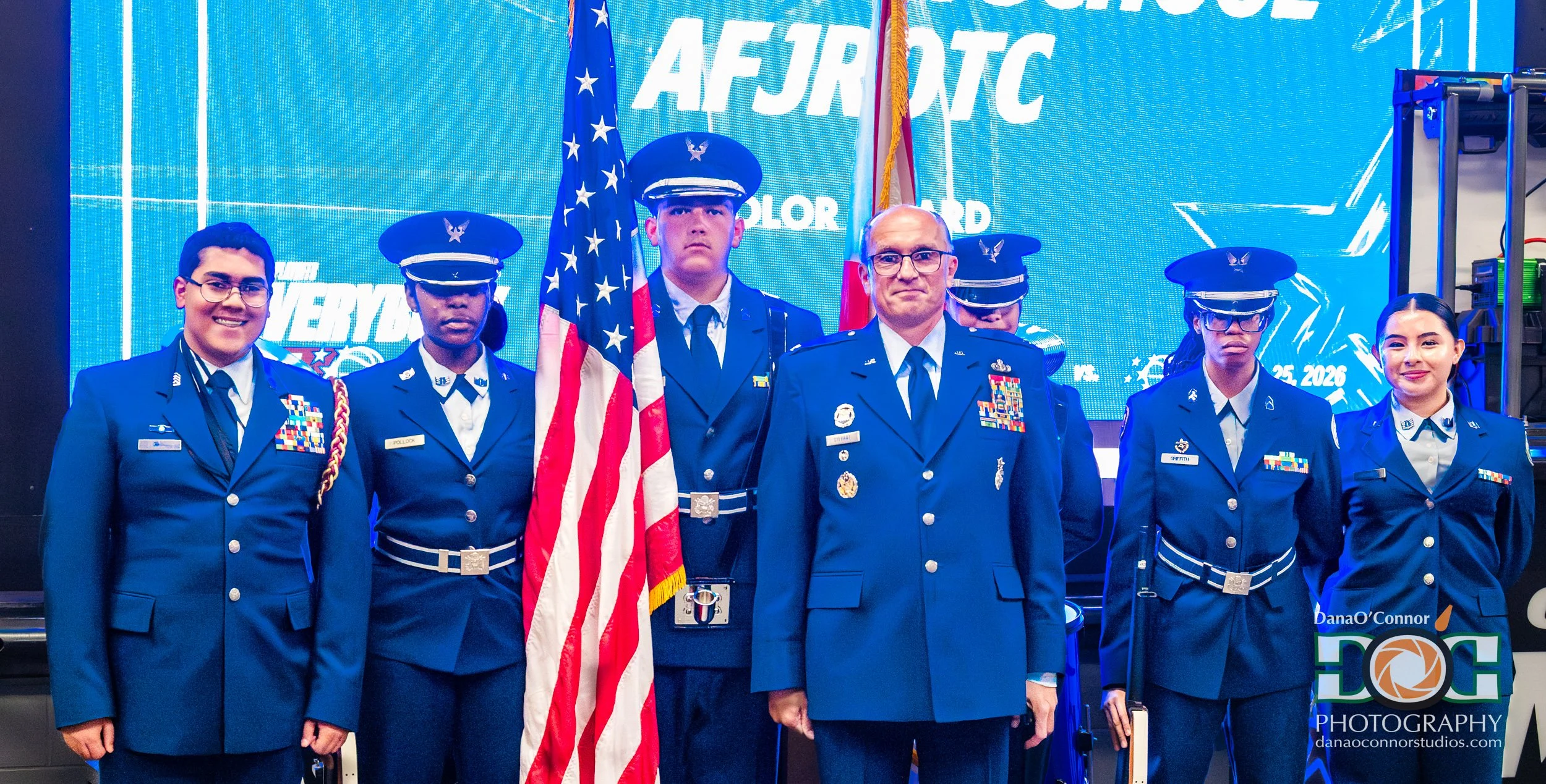 Group of people in military uniforms standing in front of a blue backdrop with white text, three of them holding flags, and two wearing glasses.