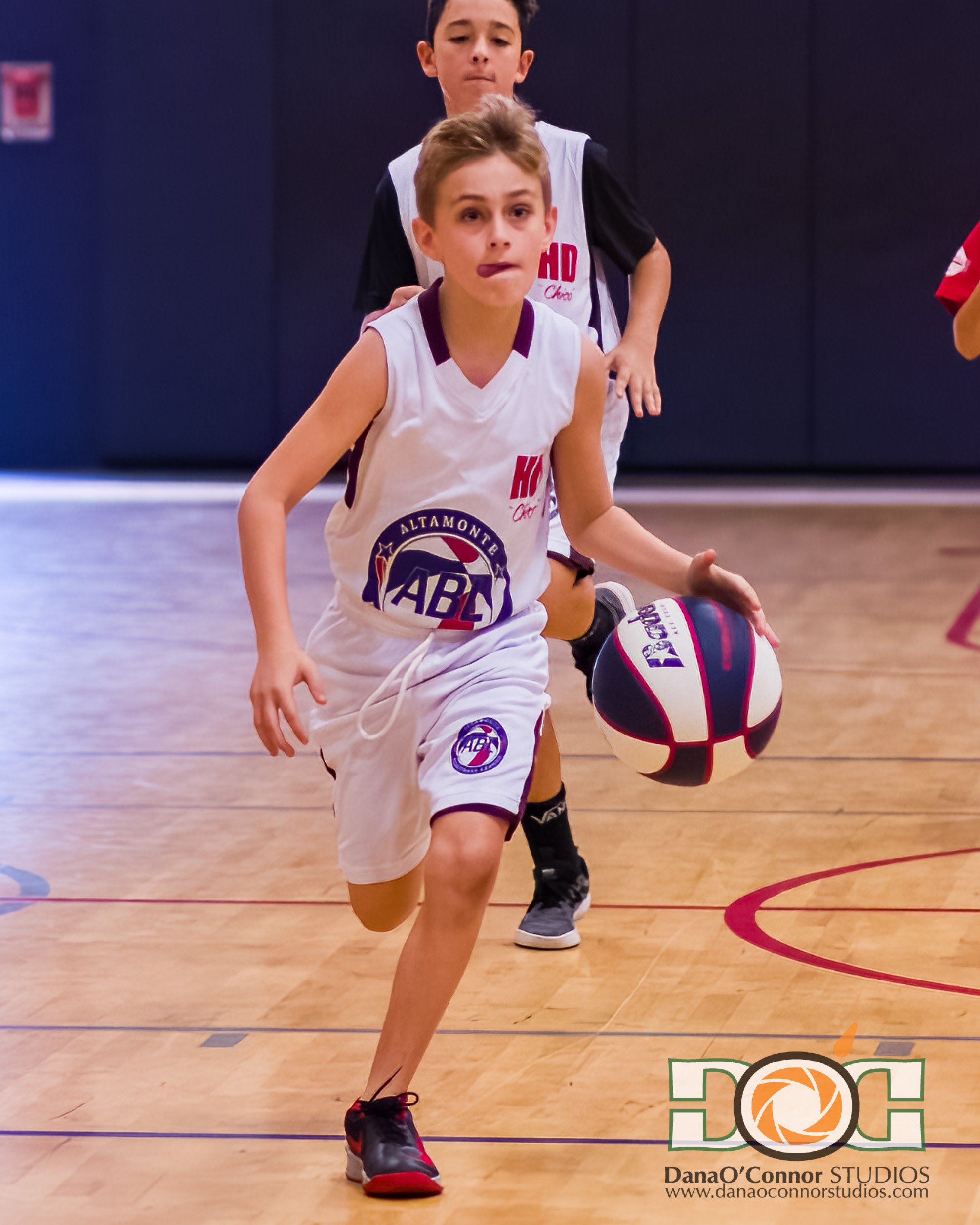Young boys playing basketball on an indoor court, with one in the foreground dribbling the ball. The gym has wooden floors and the boys are wearing youth sports uniforms.