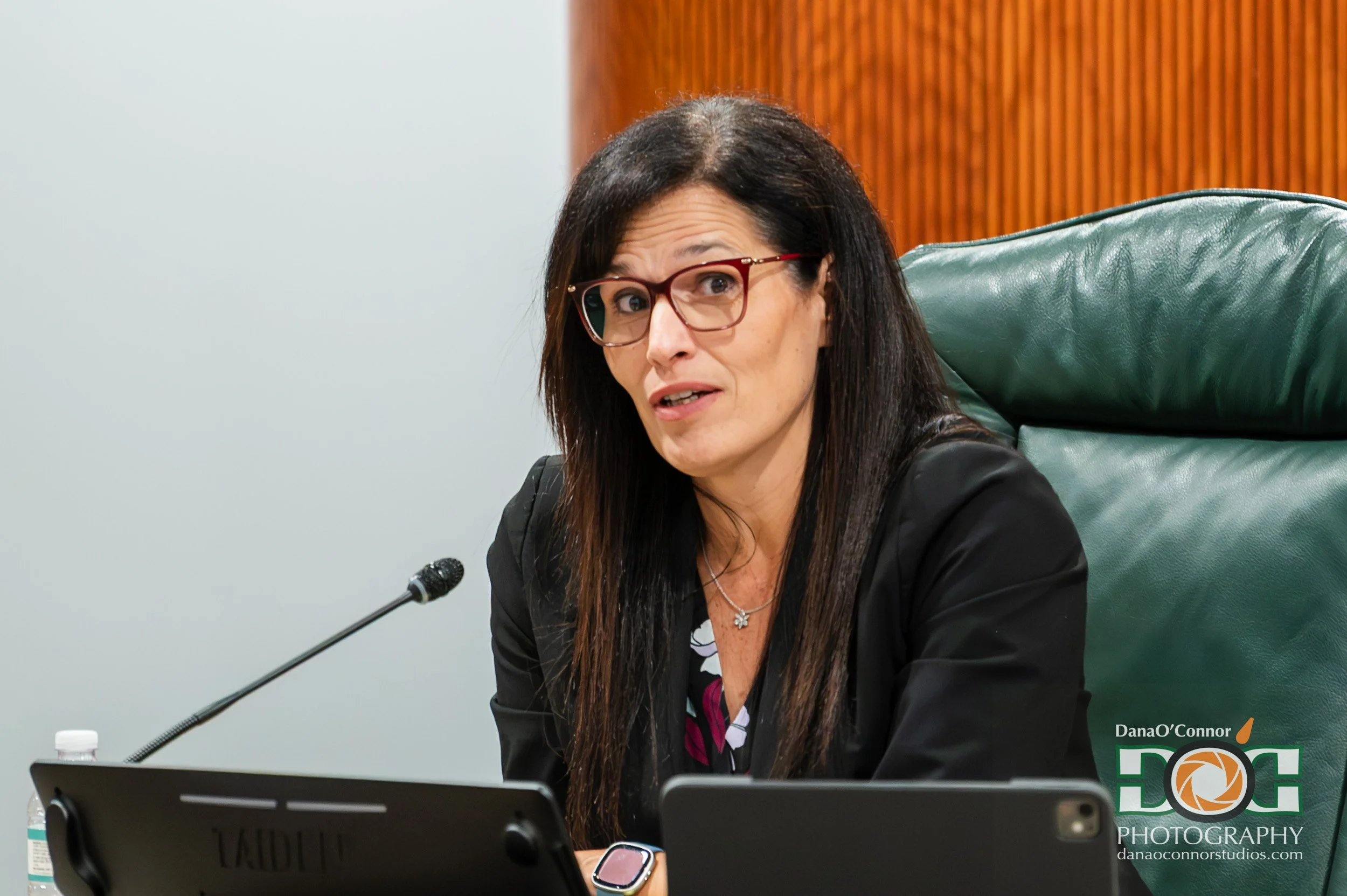 A woman with long dark hair, glasses, and a necklace, sitting at a table with a microphone and a tablet, in a conference room with wooden paneling and a green leather chair.