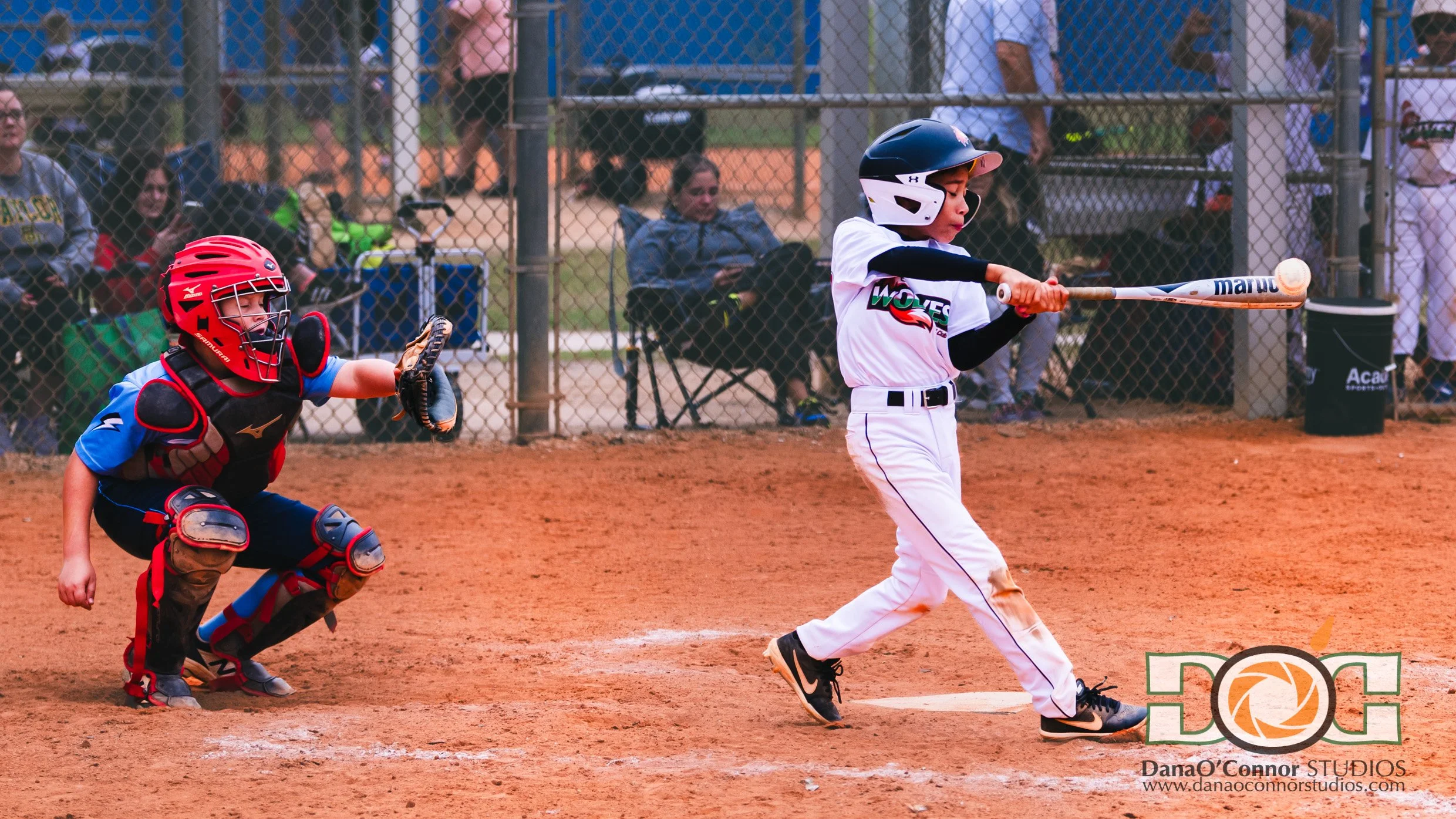 A boy in a white baseball uniform and helmet swinging a bat at a pitched ball during a baseball game, with a catcher in red and blue gear and a crowd behind a chain-link fence watching.
