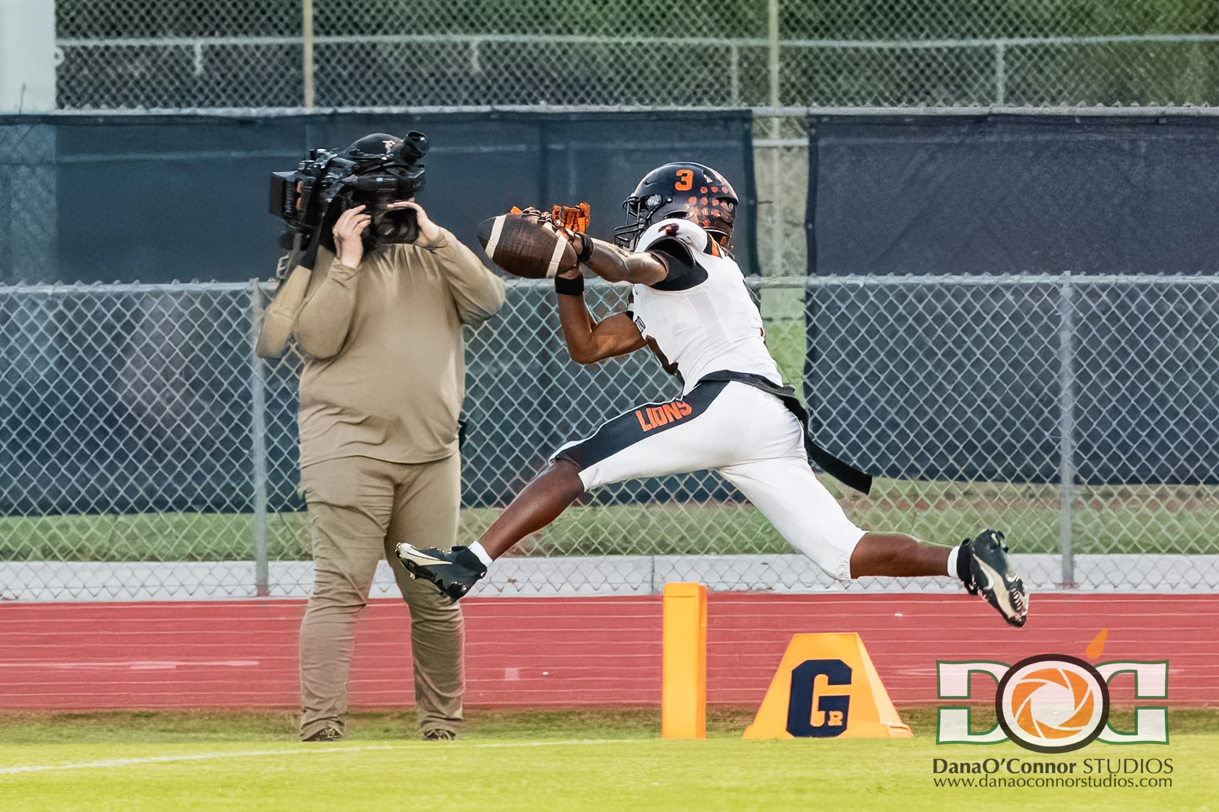 An American football player wearing a black helmet with orange dots and a white jersey with black and orange accents catches a football during a game. A videographer in a tan hoodie films the player from the side. The scene takes place on a football 