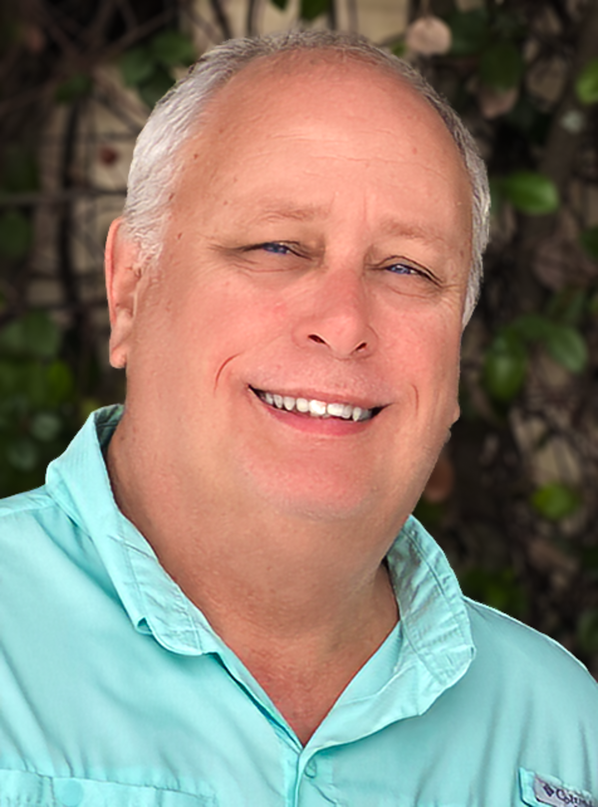 A smiling man with short, gray hair wearing a light blue shirt, standing in front of a leafy background.