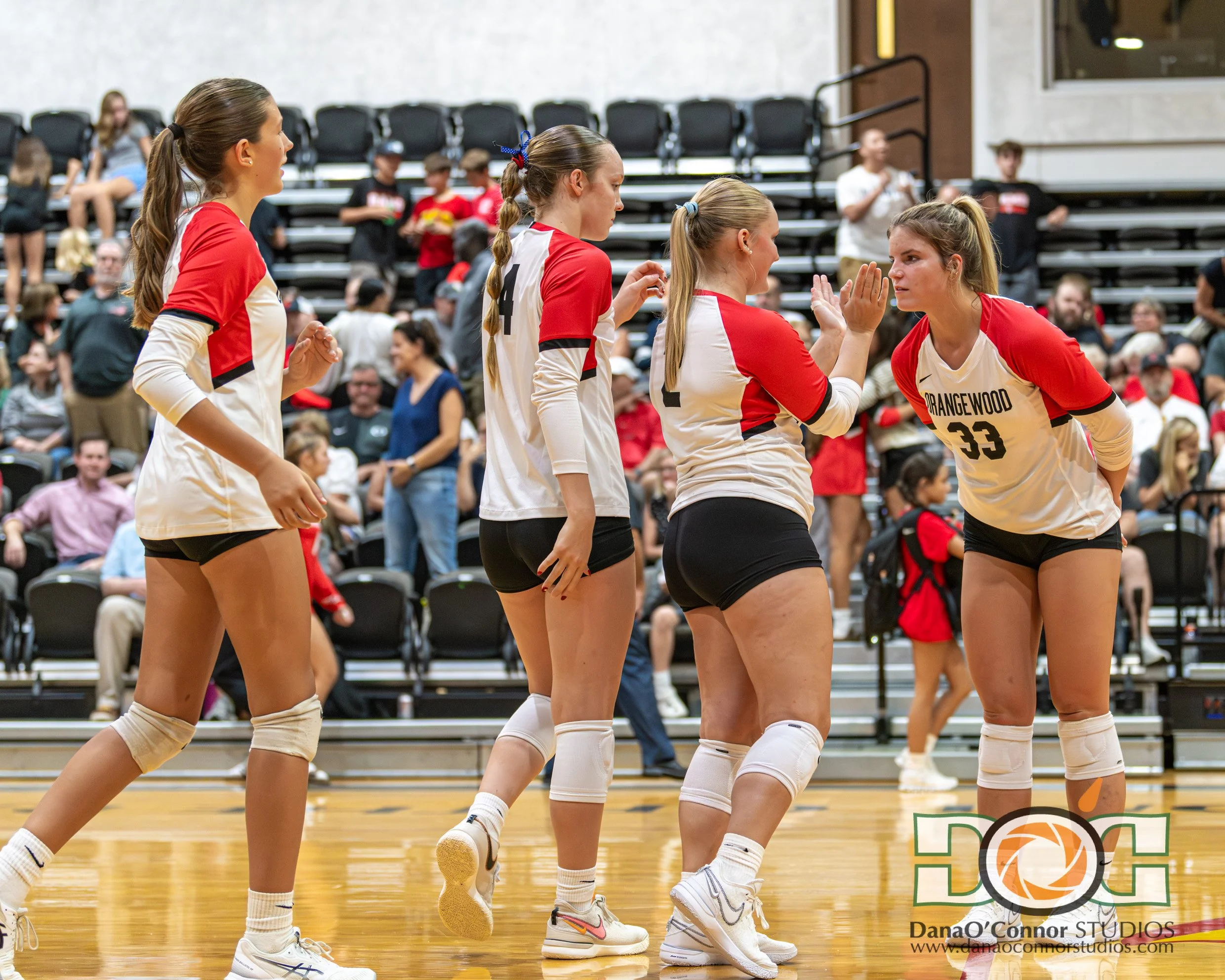 Four Orangewood Christian School's female volleyball players in red and white uniforms gather for quick meeting during a game on an indoor court, with spectators watching from the bleachers in the background.
