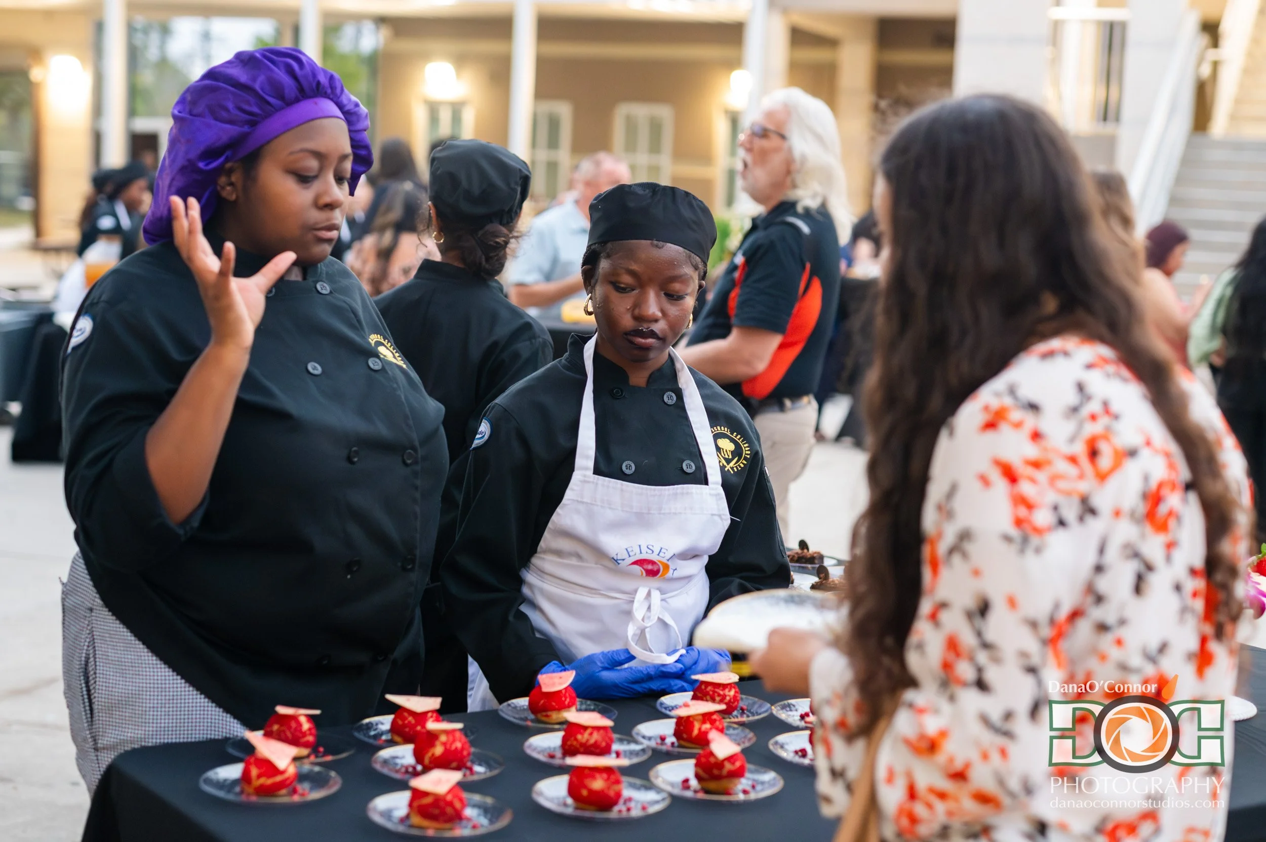 Women chefs serving desserts at an outdoor event, with a focus on a woman with purple headscarf and another with a black chef hat, while a young girl in a floral dress interacts with them.