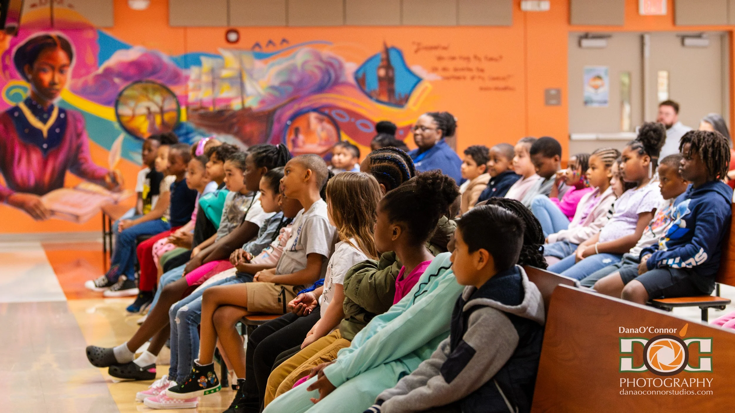 A group of children sitting in rows at an indoor event, facing forward, with colorful artwork on the orange wall behind them.